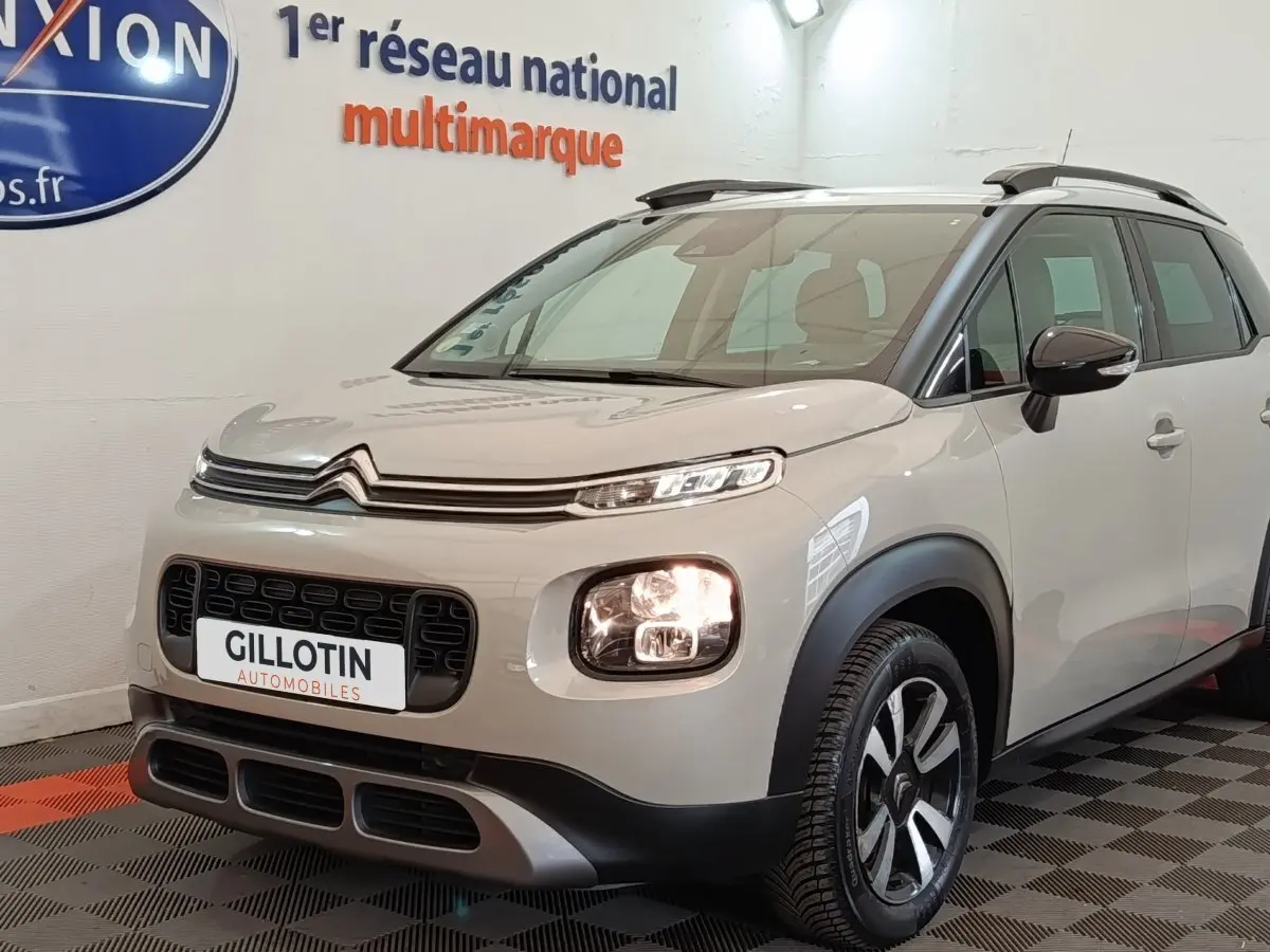 Vue 3/4 avant droite d'un Citroën C3 Aircross beige avec feux allumés et barres de toit noires en intérieur showroom.