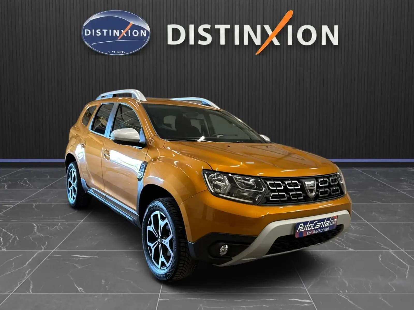 Dacia Duster orange en 3/4 avant droit, avec jantes bi-ton et barres de toit argentées, sur sol carrelé intérieur showroom.