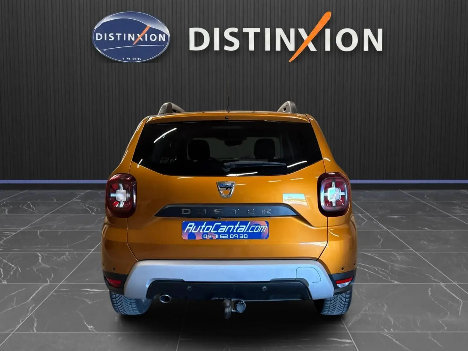 Vue arrière d'un Dacia Duster orange 2020 avec attelage, dans un showroom au sol gris et mur noir marqué Distinxion.