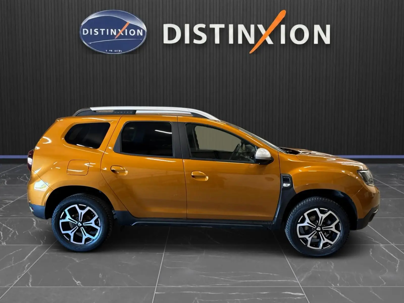 Profil droit d'un Dacia Duster orange 2020 avec jantes bi-ton et barres de toit argentées, sur fond noir Distinxion.