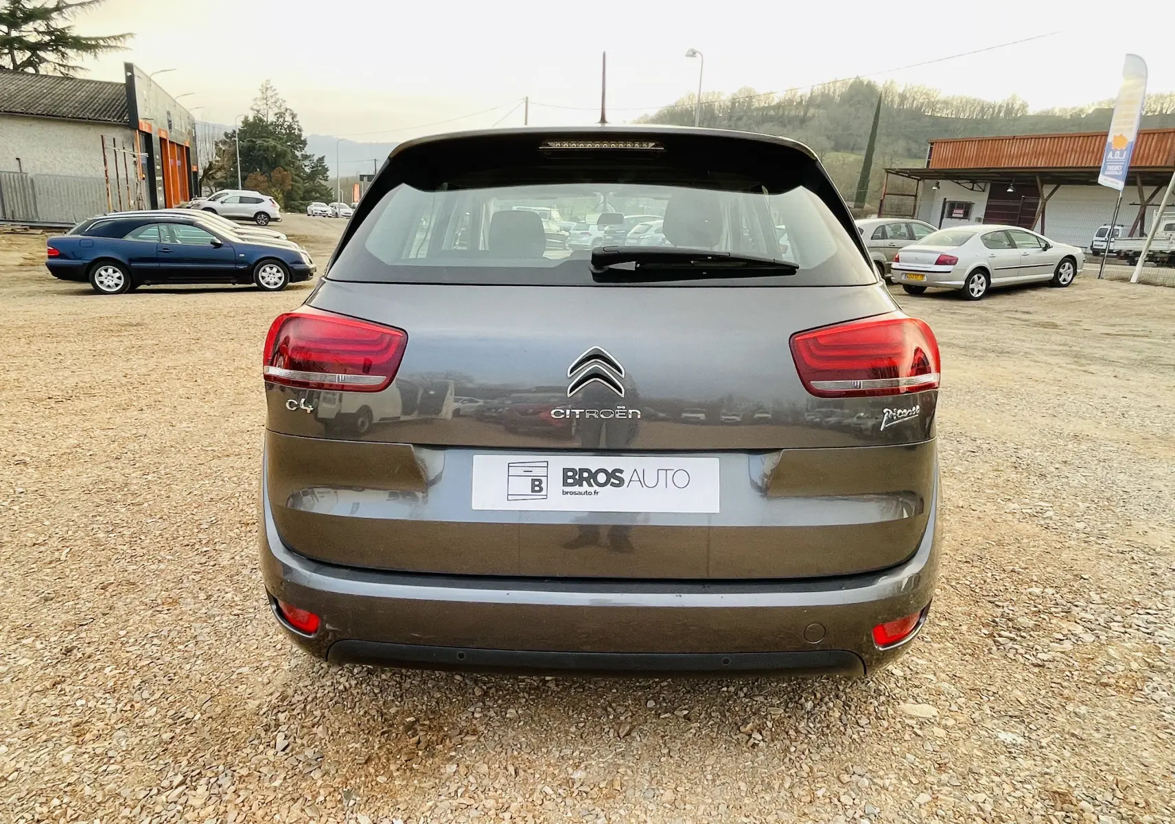 Vue arrière d'une Citroën C4 Picasso gris métal garée sur un sol en gravier avec plusieurs voitures en arrière-plan.