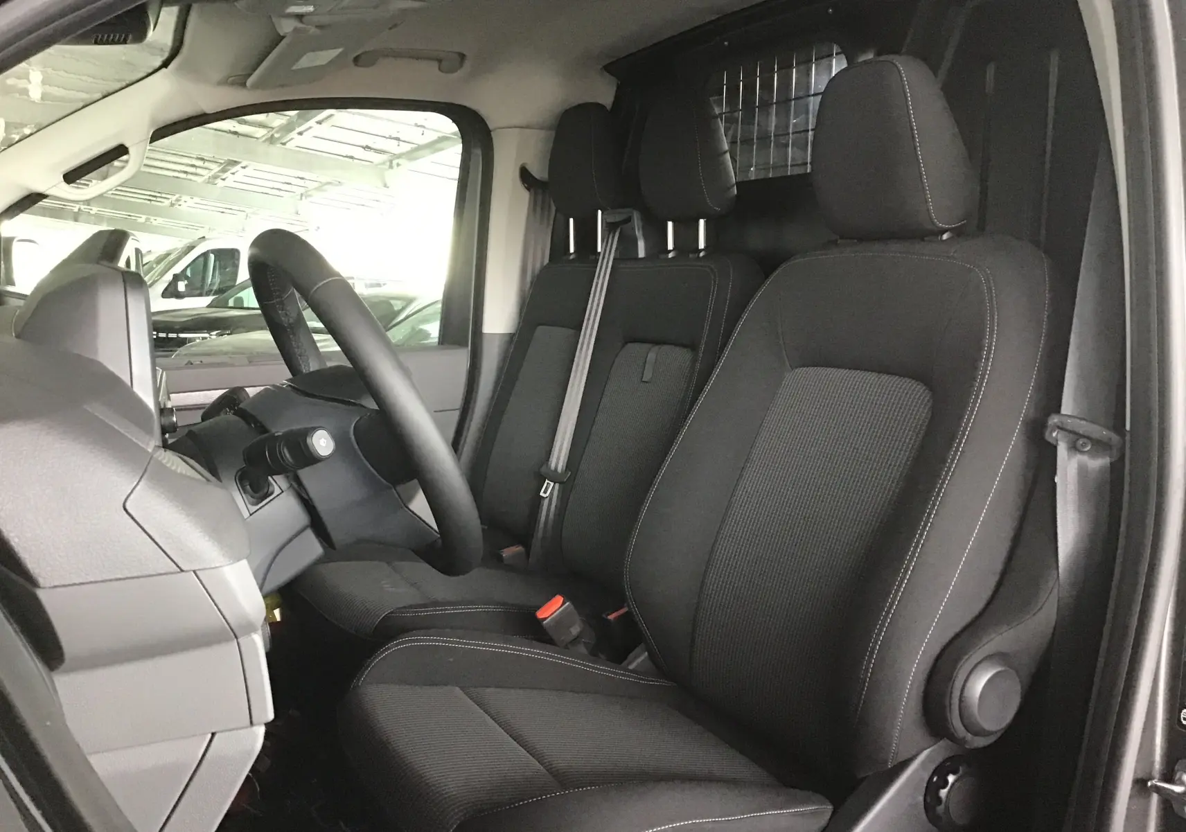 Vue intérieure côté conducteur du Volkswagen Transporter T7 avec sièges noirs et séparation métallique derrière.