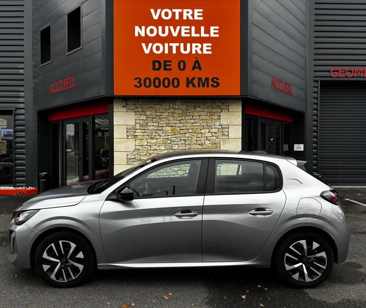 Profil côté gauche d'une Peugeot 208 PureTech 100 S&S Active gris clair, garée devant un bâtiment avec panneau orange.