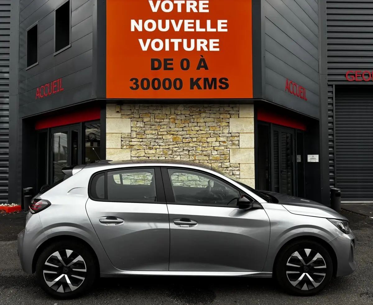 Profil latéral droit d'une Peugeot 208 gris clair garée devant un bâtiment avec enseigne orange et murs en pierre.