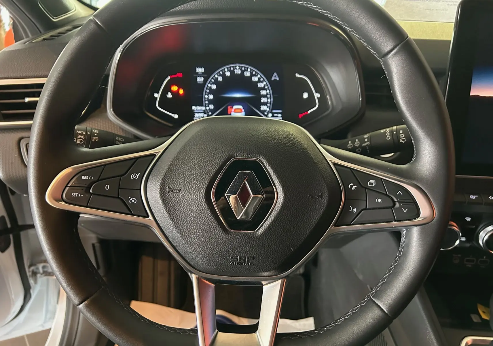 Vue rapprochée du volant en cuir noir avec logo Renault et tableau de bord numérique éclairé d'une Clio V blanche nacrée.