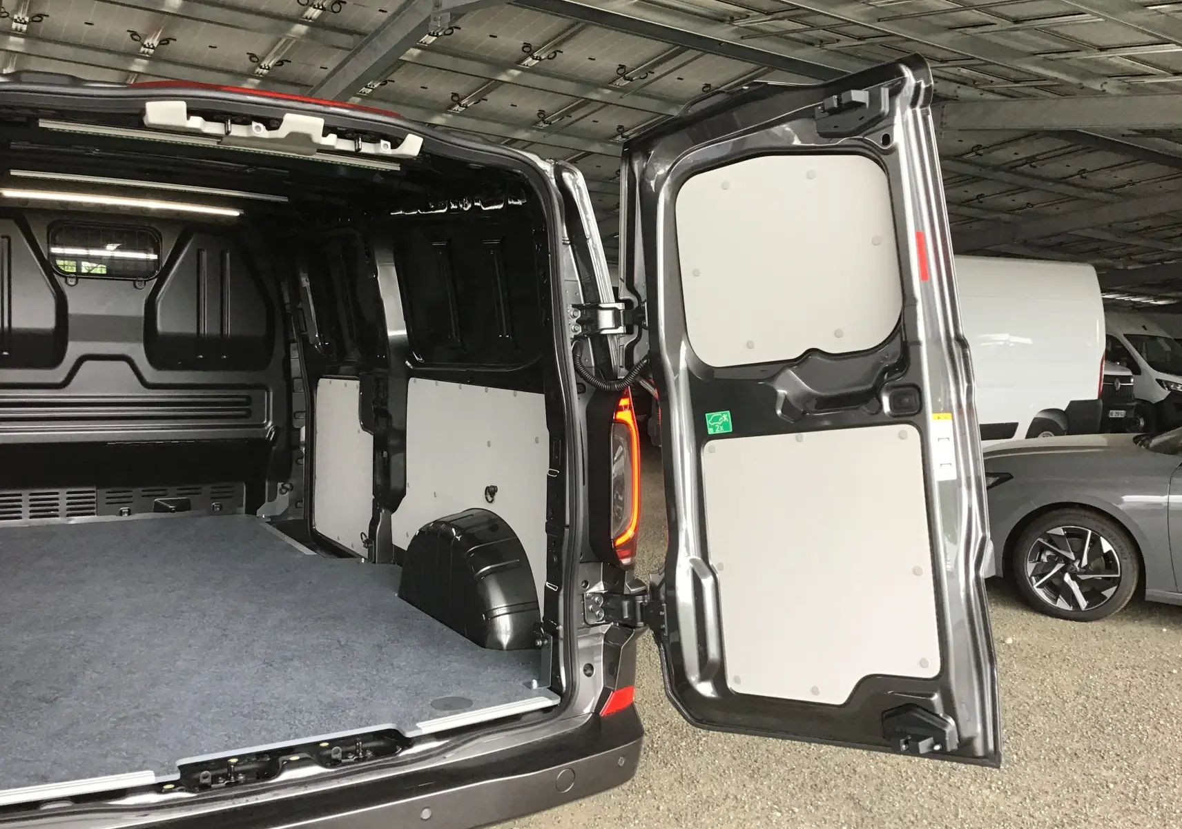 Vue arrière du Volkswagen Transporter T7 gris anthracite avec portes ouvertes dévoilant l'espace de chargement vide et les panneaux intérieurs clairs.