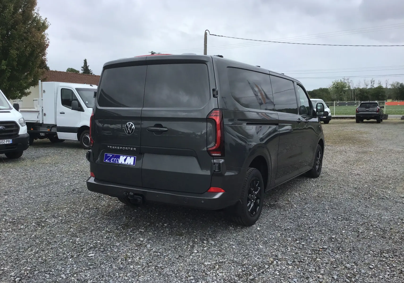 Vue 3/4 arrière droite d'un Volkswagen Transporter T7 gris anthracite avec barres de toit repliables sur un parking.