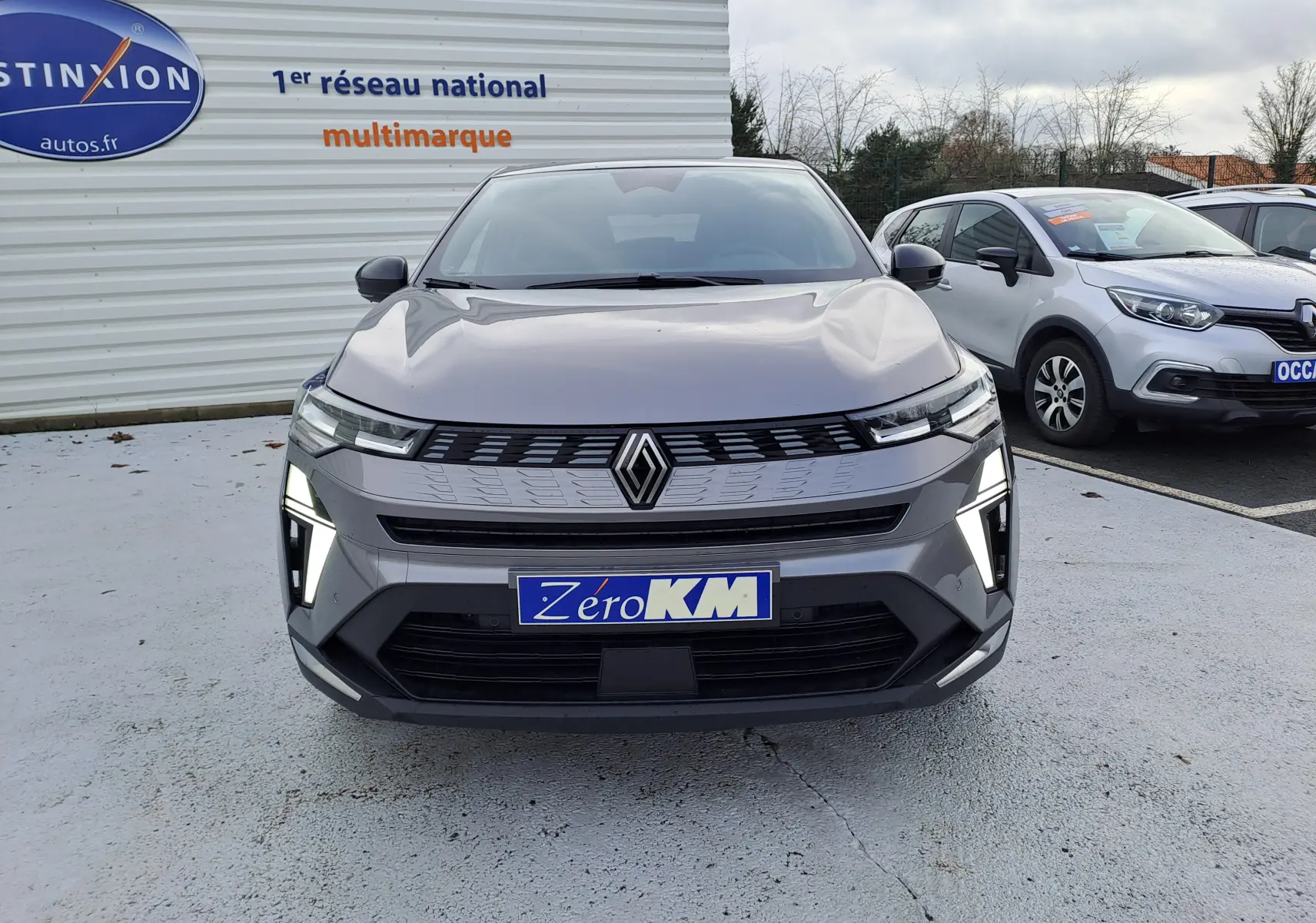 Vue de face d'une Renault Symbioz gris cassiopée avec feux LED allumés sur un parking extérieur.
