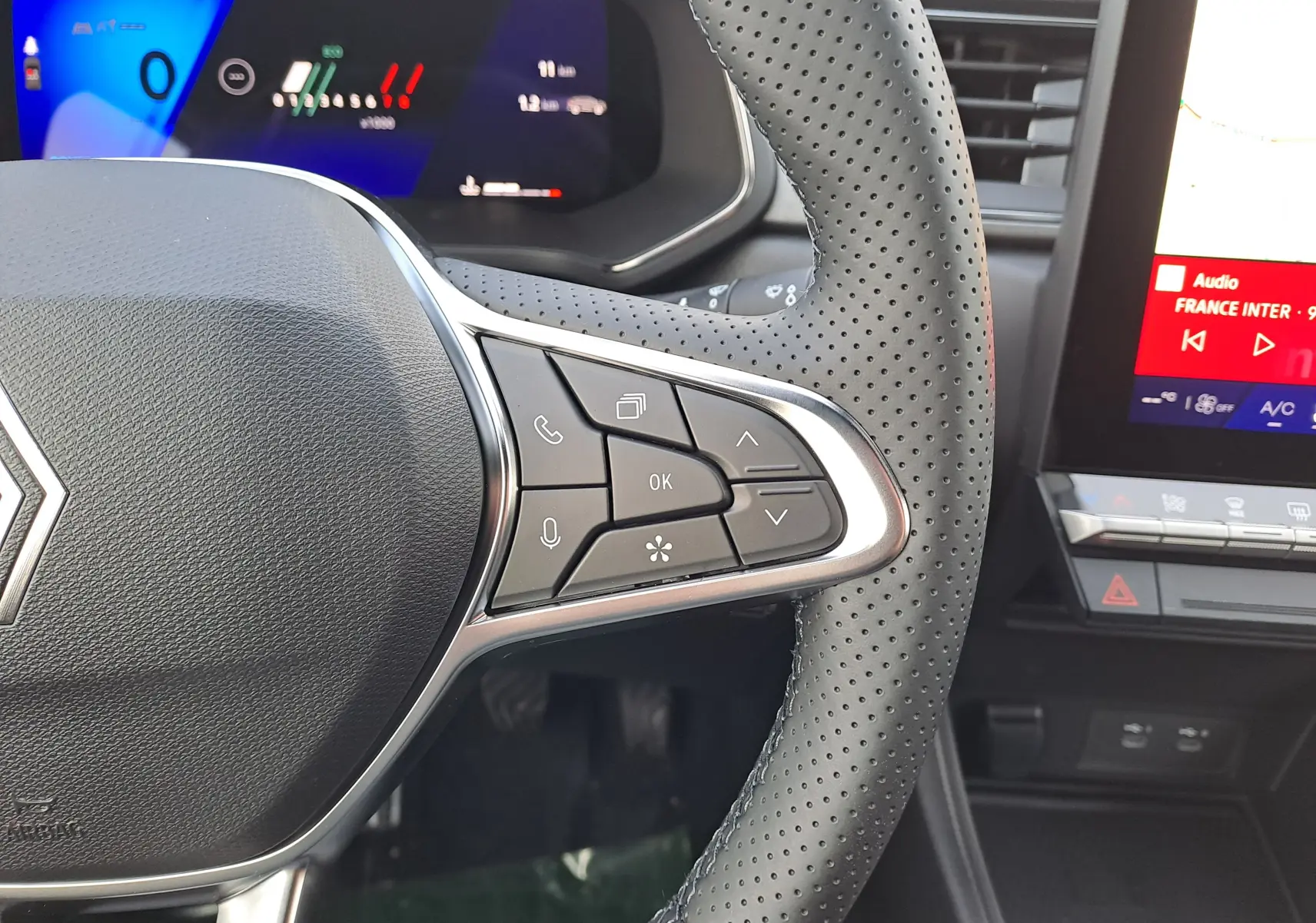 Gros plan sur le volant perforé gris cassiopée du Renault Symbioz 2025, avec commandes intégrées et tableau de bord numérique en arrière-plan.