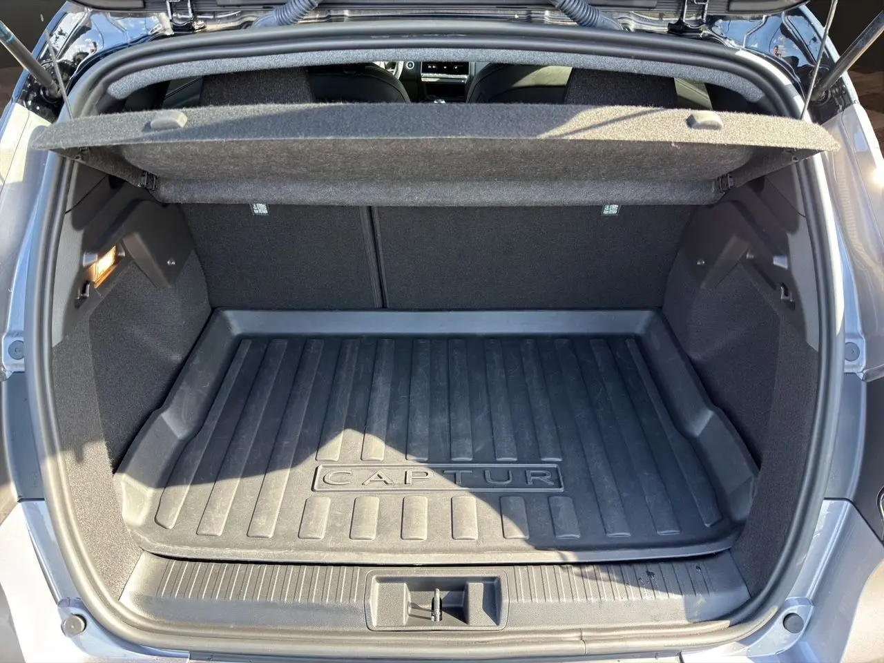 Coffre ouvert vu de l'arrière d'un Renault Captur gris Cassiopée avec tapis de coffre noir marqué Captur.