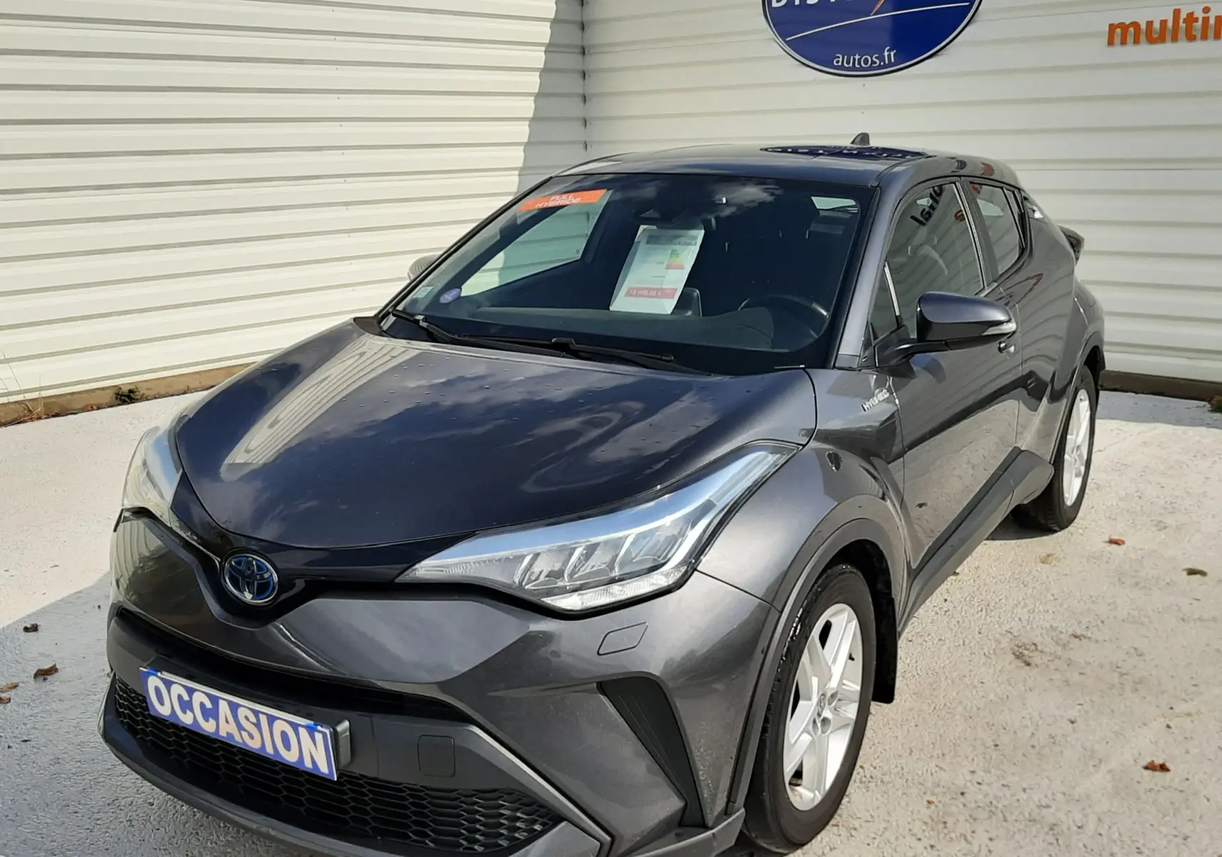 Toyota C-HR 1.8 Hybrid gris atlas métal vu en 3/4 avant droit, avec plaque "OCCASION" et logo Distinxion en arrière-plan.