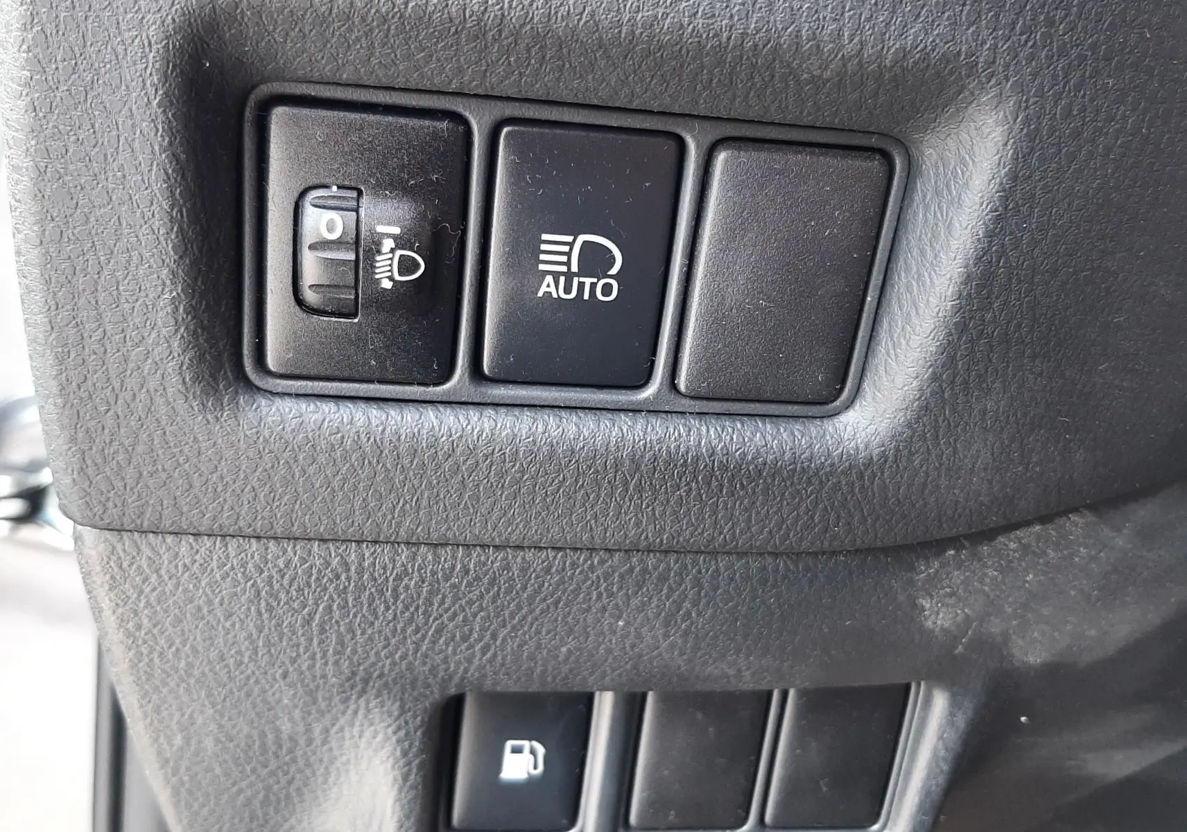 Gros plan sur les boutons de réglage des phares et de l'ouverture du réservoir, intérieur gris du Toyota C-HR 1.8 Hybrid.