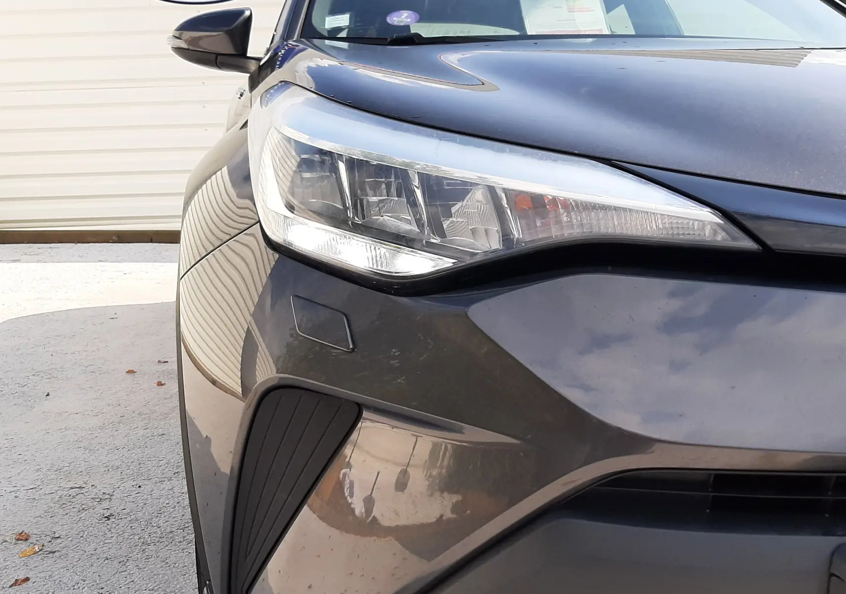 Gros plan sur l'avant gauche du Toyota C-HR 1.8 Hybrid gris atlas métal, mettant en valeur le phare et la calandre.
