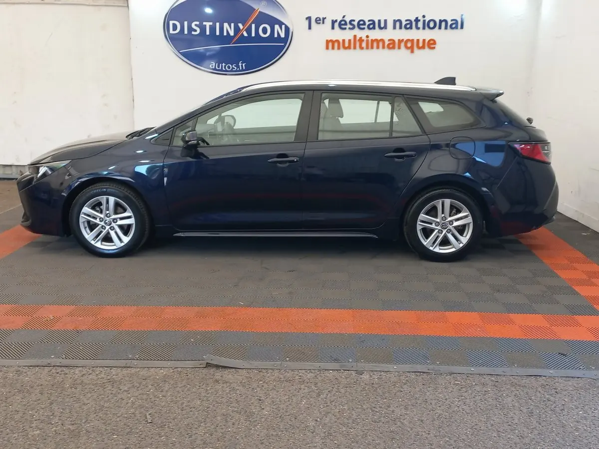 Vue latérale gauche d'une Toyota Corolla bleue hybride 2022 en version break, stationnée en intérieur showroom.