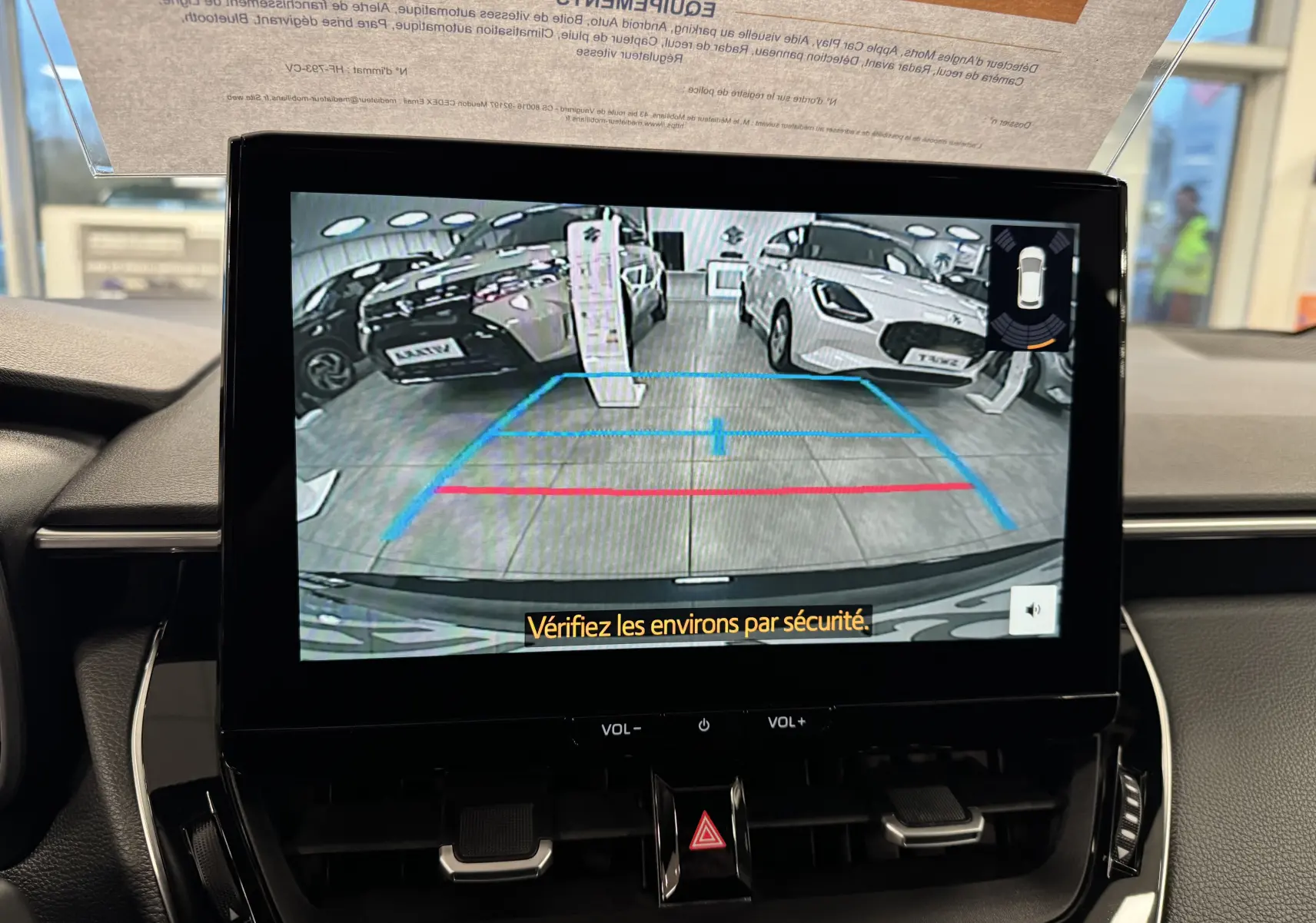 Vue de l'écran intérieur affichant la caméra de recul avec deux voitures en exposition, dont une Suzuki Swace blanche à droite.