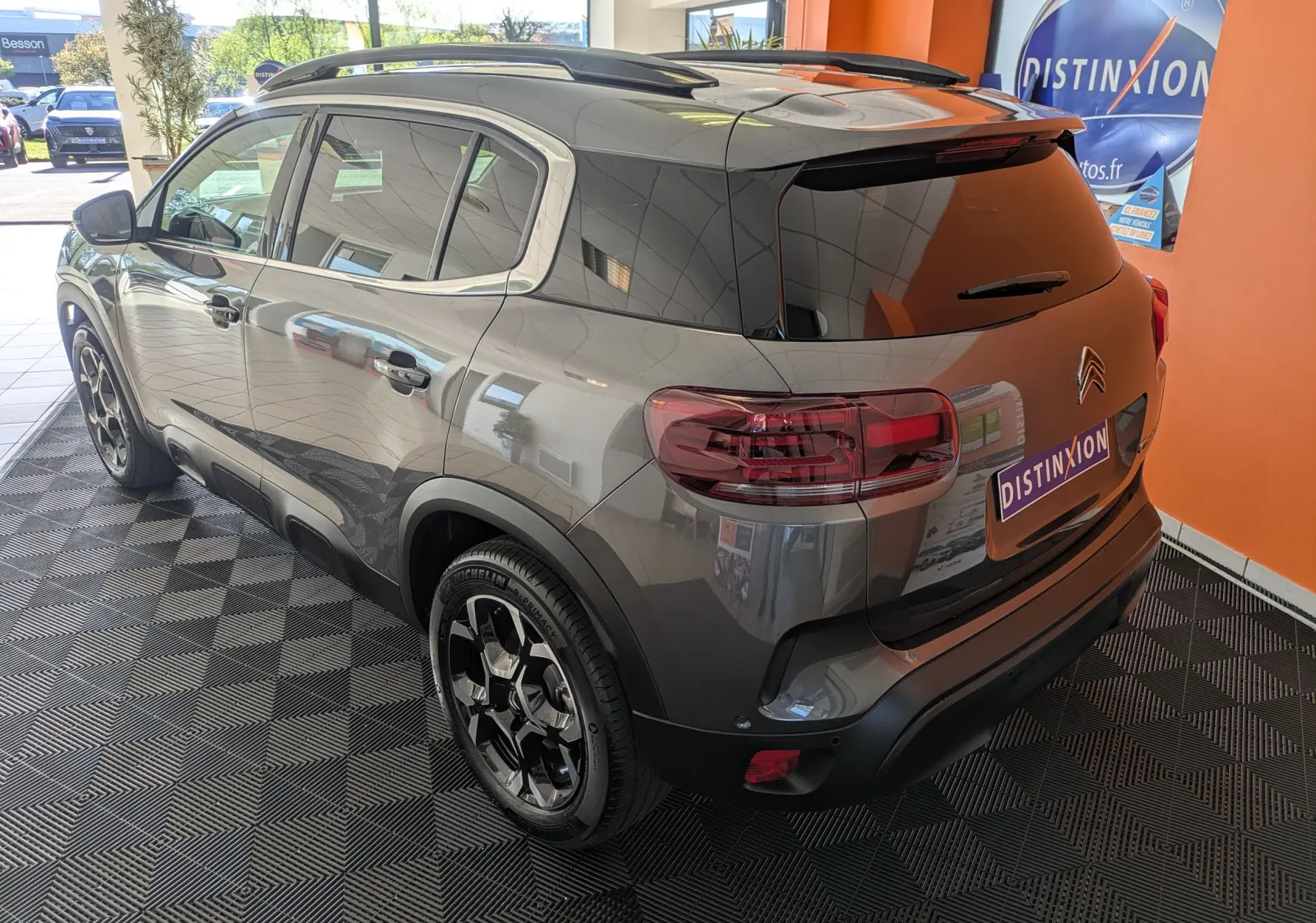 Vue 3/4 arrière droit du Citroën C5 Aircross gris platinium avec jantes biton 18 pouces et barres de toit visibles en intérieur showroom.