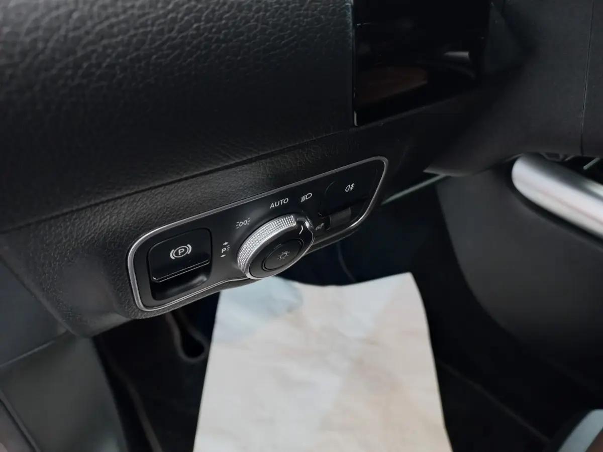 Gros plan sur le panneau de commande d’éclairage et frein à main électrique côté gauche intérieur d’une Mercedes GLA gris foncé.