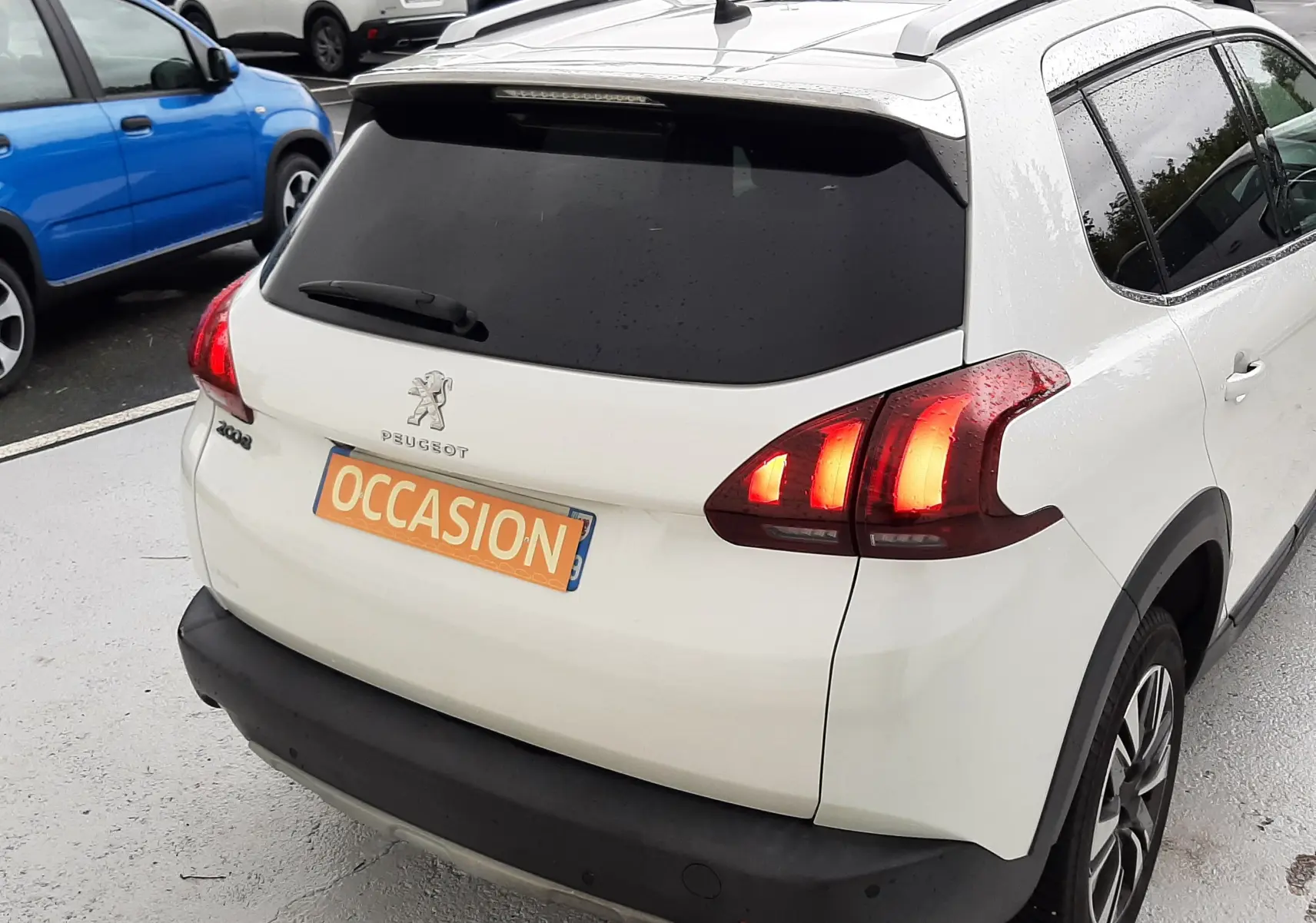 Vue 3/4 arrière droite d'un Peugeot 2008 blanc de 2017 avec feux arrière allumés et plaque "OCCASION" visible.