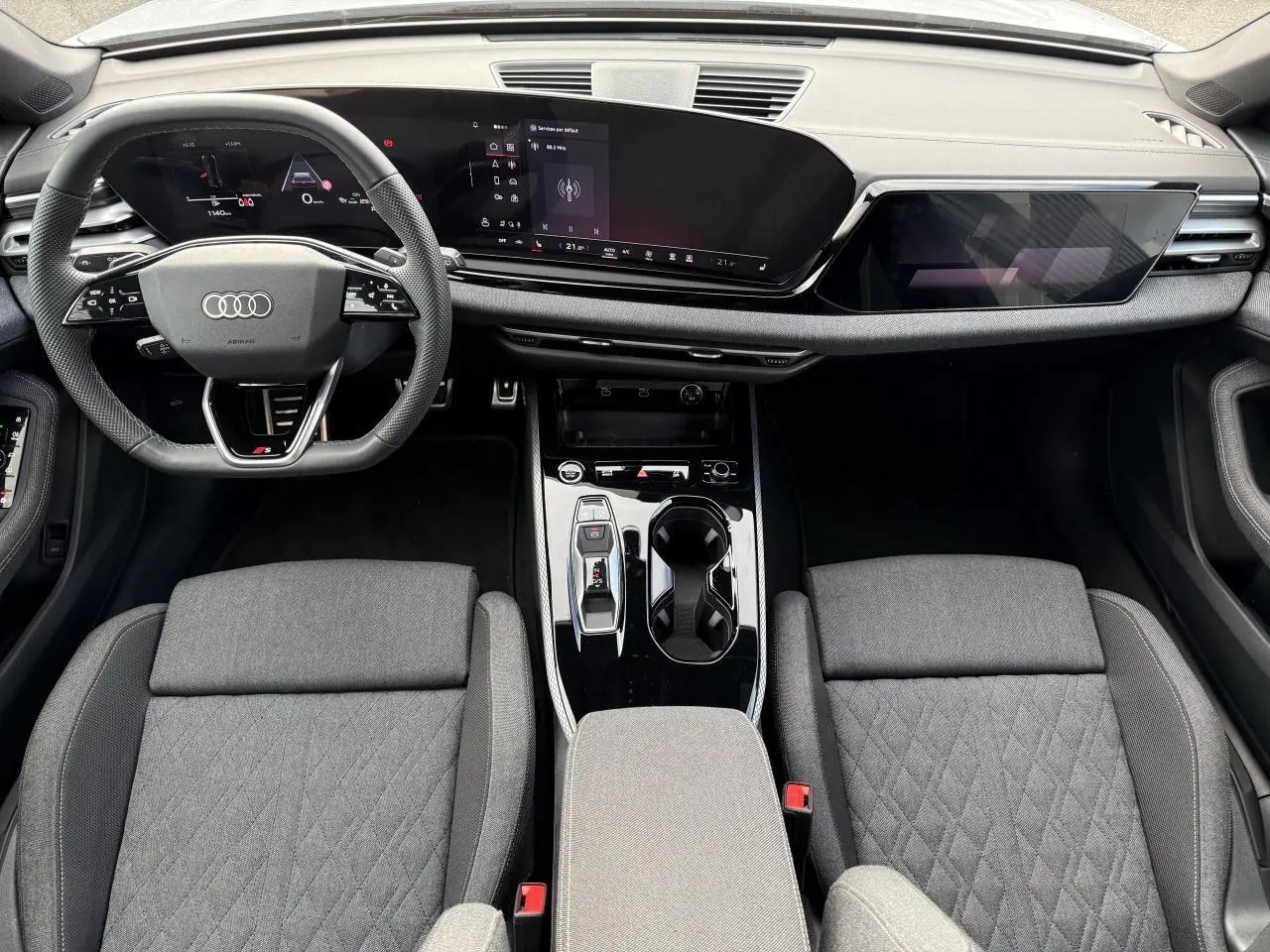 Vue intérieure de l'Audi A5 2025 montrant le tableau de bord numérique et les sièges tissu gris avec console centrale noire brillante.