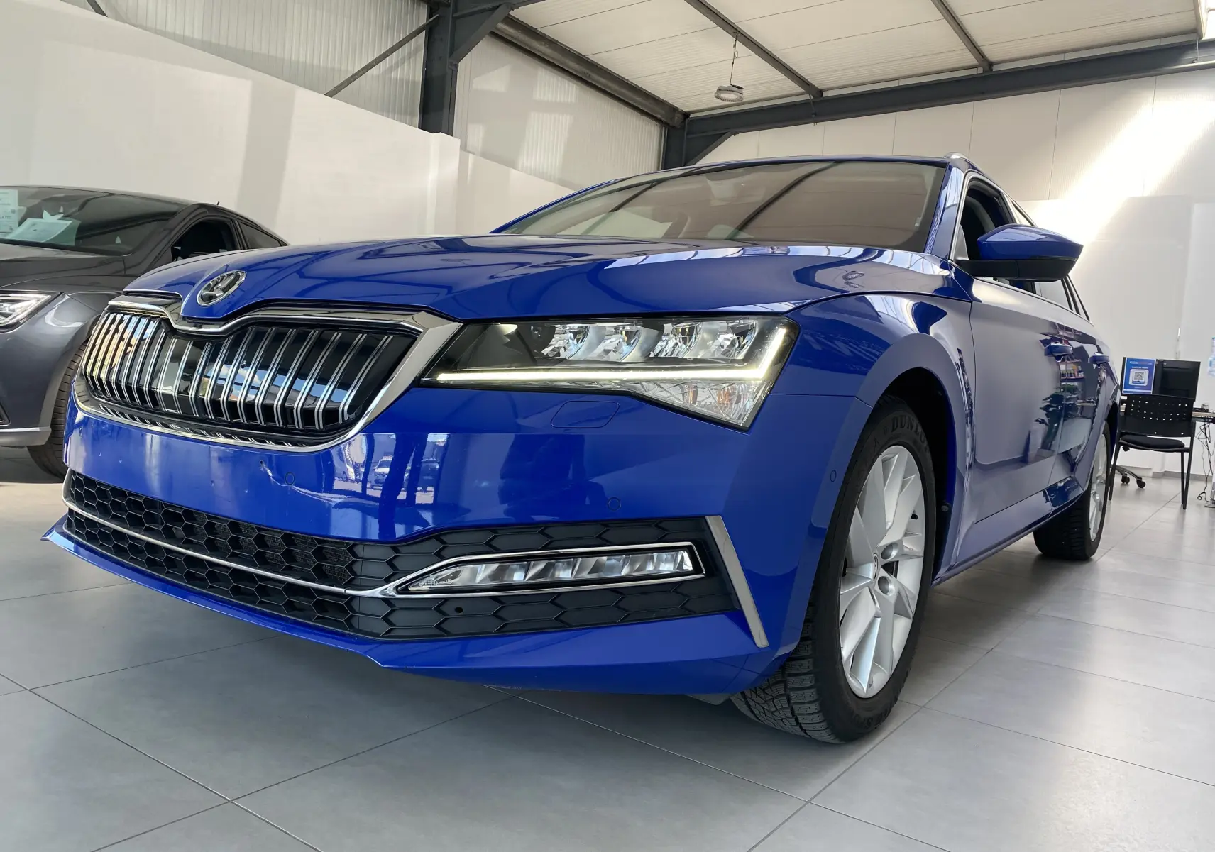 Vue 3/4 avant droite d'une Skoda Superb Combi bleu énergie avec calandre chromée et phares LED allumés dans un showroom.