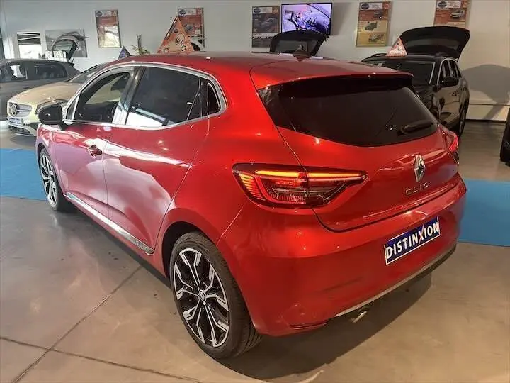 Vue 3/4 arrière droite d'une Renault Clio V rouge flamme métal avec feux arrière allumés en showroom.