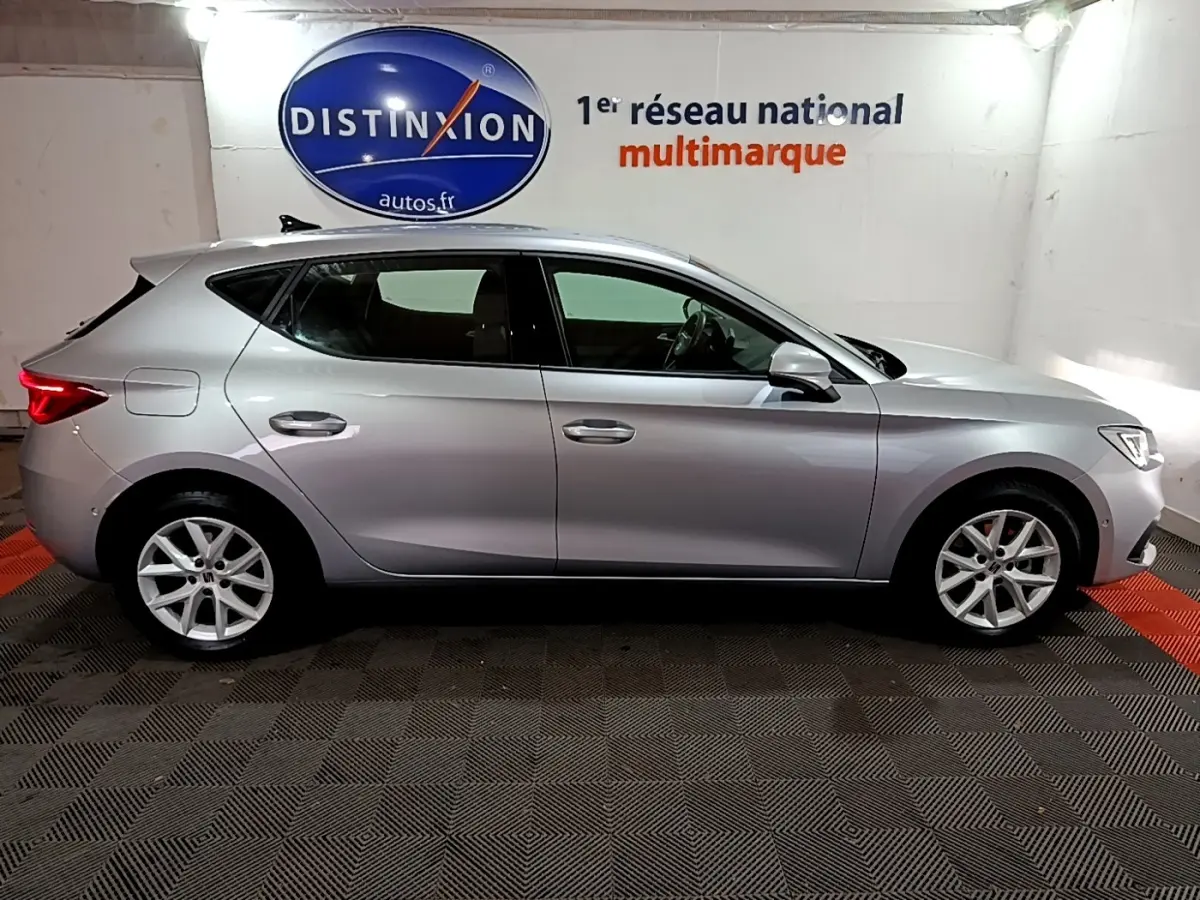 Profil côté droit d'une SEAT LEON gris clair 2023 dans un showroom avec logo Distinxion en arrière-plan.