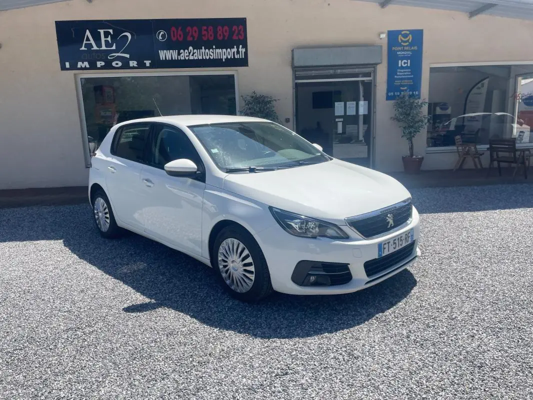 PEUGEOT 308 blanc vue 3/4 avant droit, garée devant un bâtiment avec panneaux publicitaires visibles.