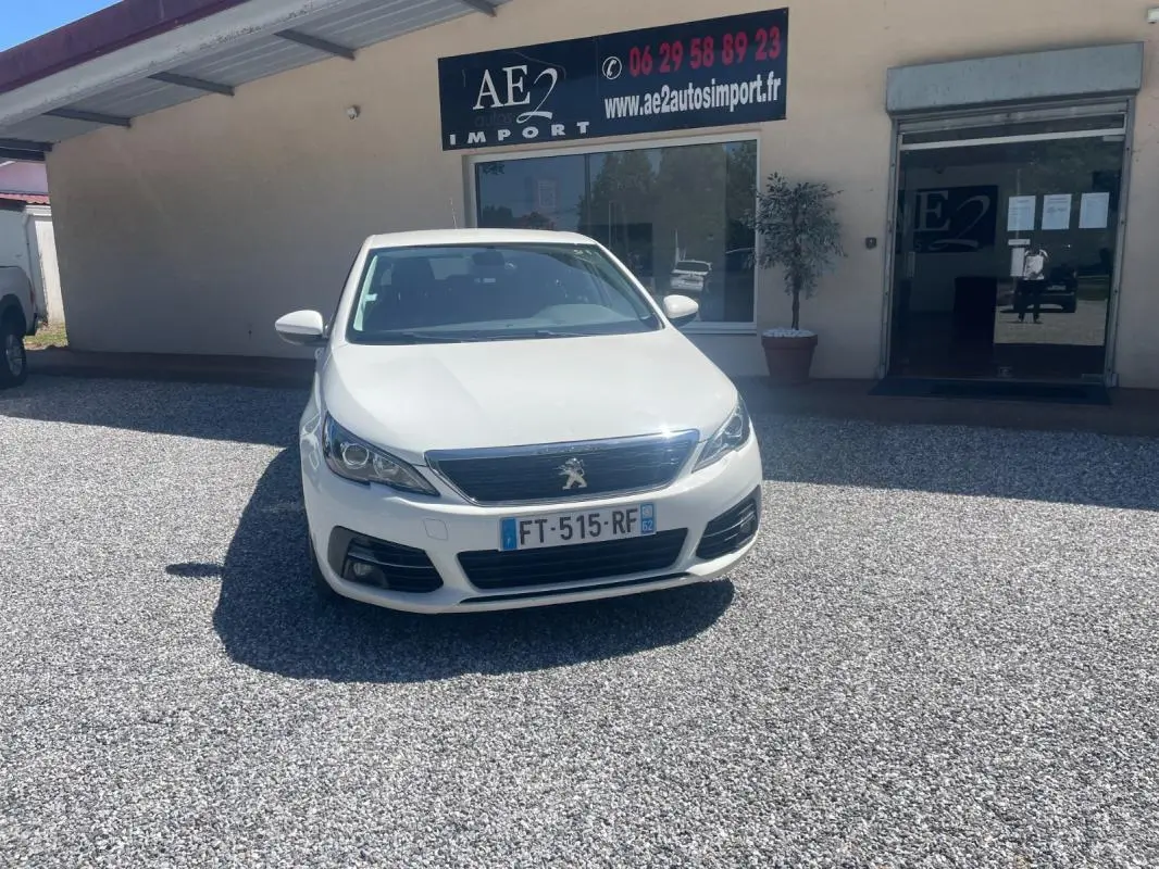 Peugeot 308 blanc vue de face, stationnée devant un bâtiment avec panneau AE2 Import visible en arrière-plan.