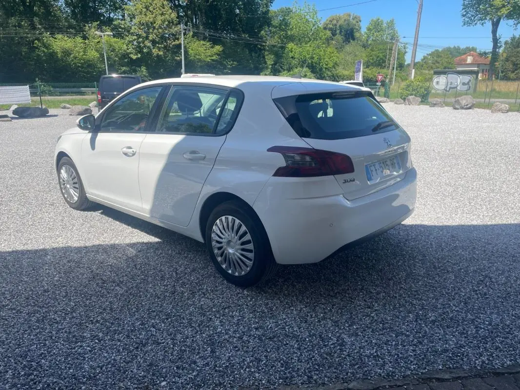 Peugeot 308 blanc vue 3/4 arrière côté gauche sur parking gravillonné, avec feux arrière foncés et jantes spécifiques.