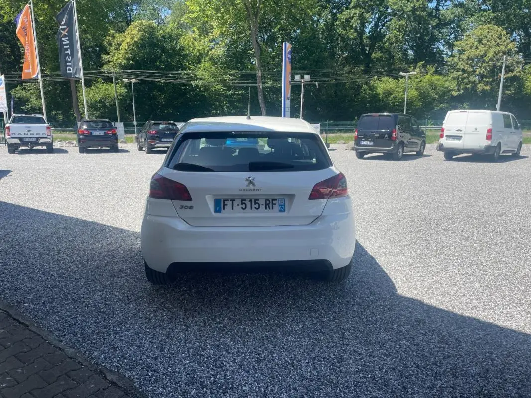 Vue arrière d'une Peugeot 308 blanche de 2020, stationnée sur un parking en gravier, avec plaques d'immatriculation visibles.