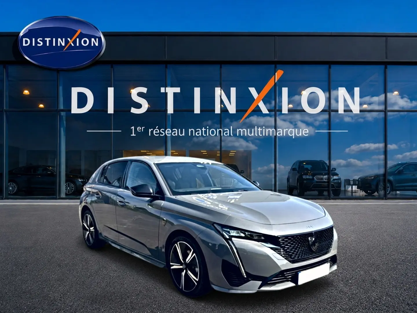 Peugeot 308 gris en 3/4 avant droit, avec calandre noire et jantes alliage distinctives, devant showroom Distinxion.