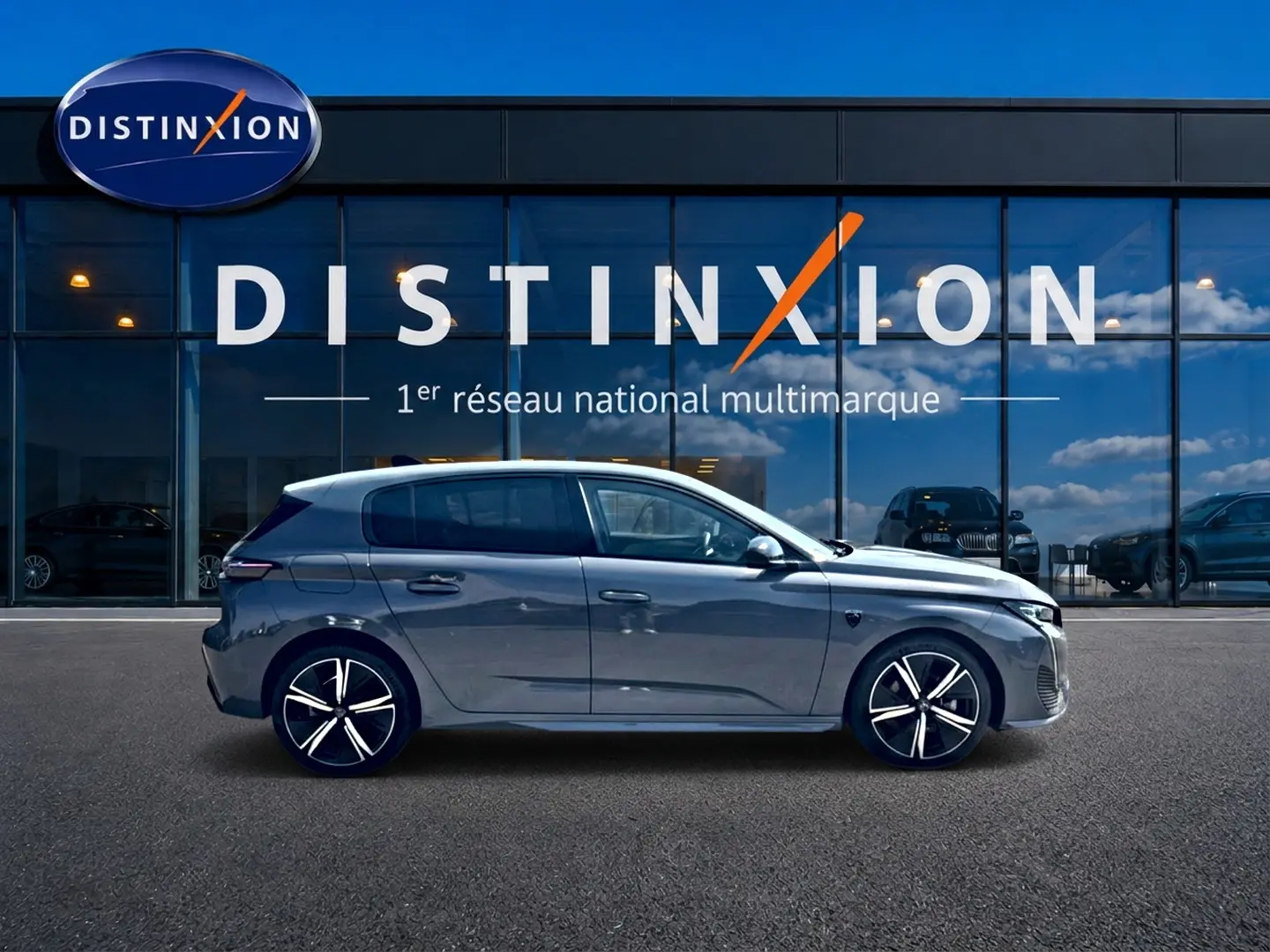 Peugeot 308 gris vue de profil côté gauche devant une vitrine avec logo Distinxion, jantes alu noires et argent.
