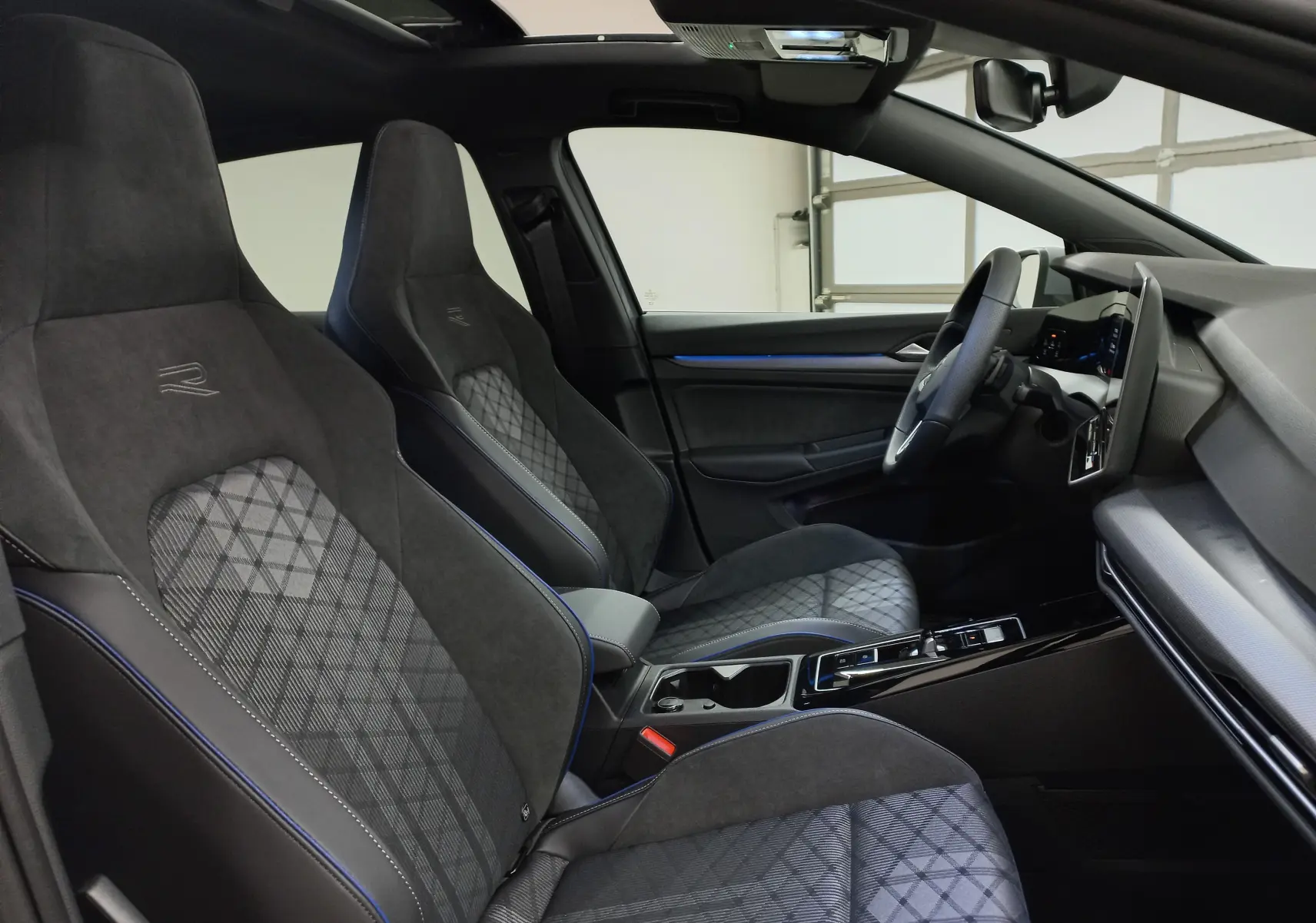 Vue intérieure côté conducteur de la Volkswagen Golf 2.0 TDI R-Line, sièges en tissu noir avec surpiqûres bleues et toit ouvrant.