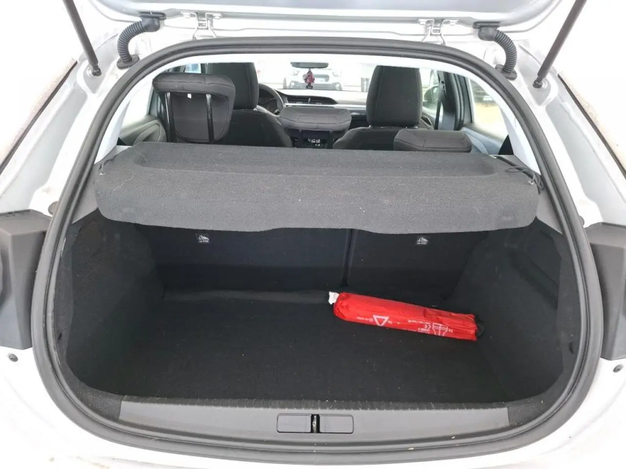 Vue arrière du coffre ouvert d'une Opel Corsa gris clair, avec cache-bagages et un triangle de sécurité rouge à l'intérieur.