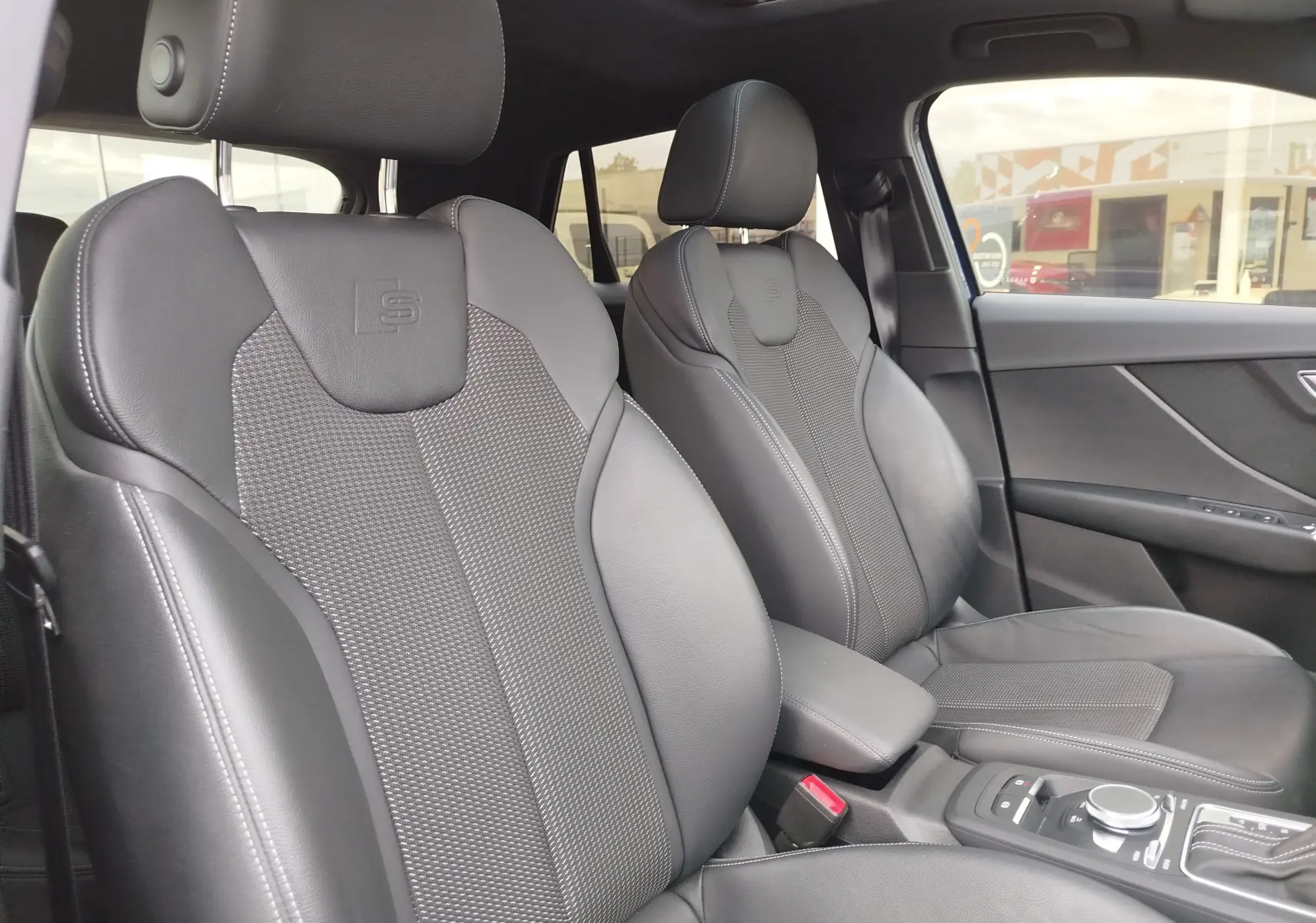 Vue intérieure de l'Audi Q2 2023, sièges avant en cuir noir avec logo S embossé et toit ouvrant panoramique.