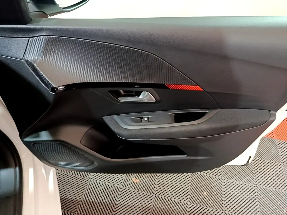 Détail de la porte avant gauche noire avec insert carbone et bande rouge d’une Peugeot 208 électrique blanche vue de l’intérieur.