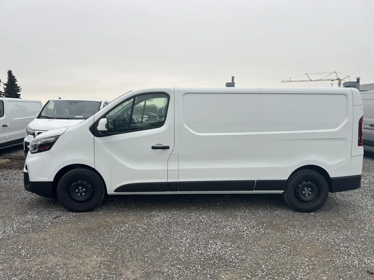Vue de profil côté gauche d’un utilitaire Nissan Primastar blanc L2H1 avec roues noires sur un sol gravillonné.