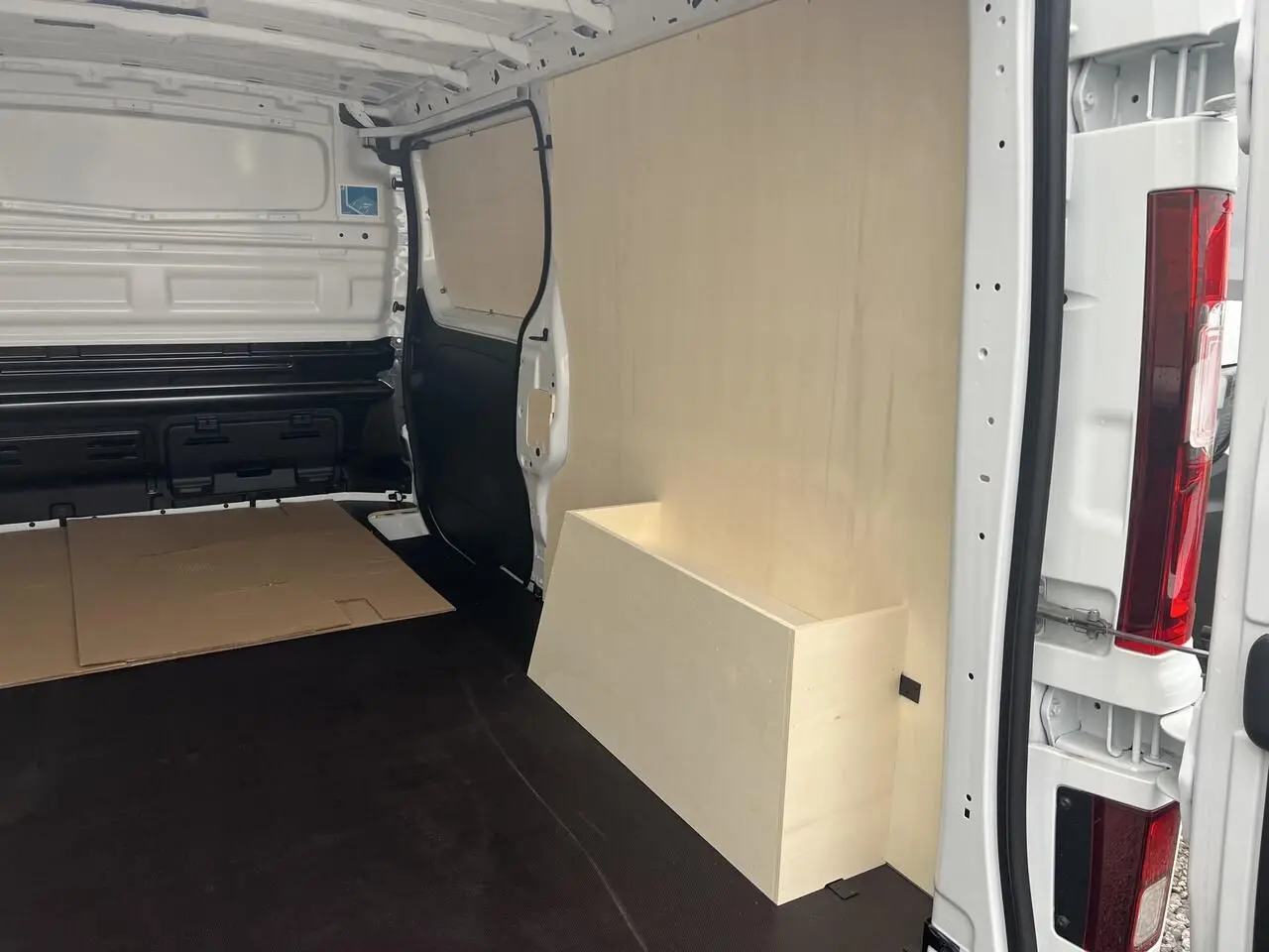 Vue intérieure arrière droite du Nissan Primastar blanc 2025 avec aménagement bois Pack Cargo et plancher noir.