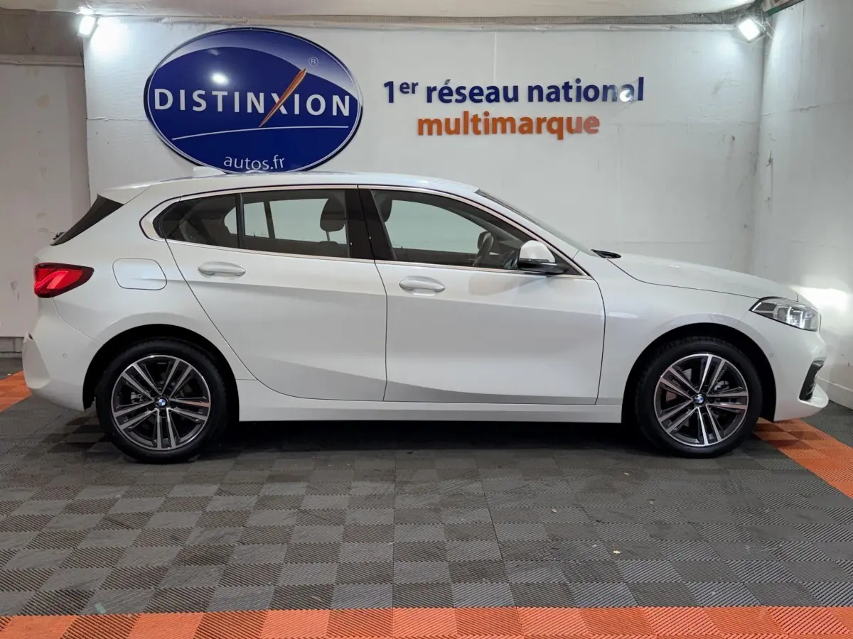 Profil côté gauche d'une BMW Série 1 116i Business Design blanche, avec jantes alliage et feux arrière LED visibles.