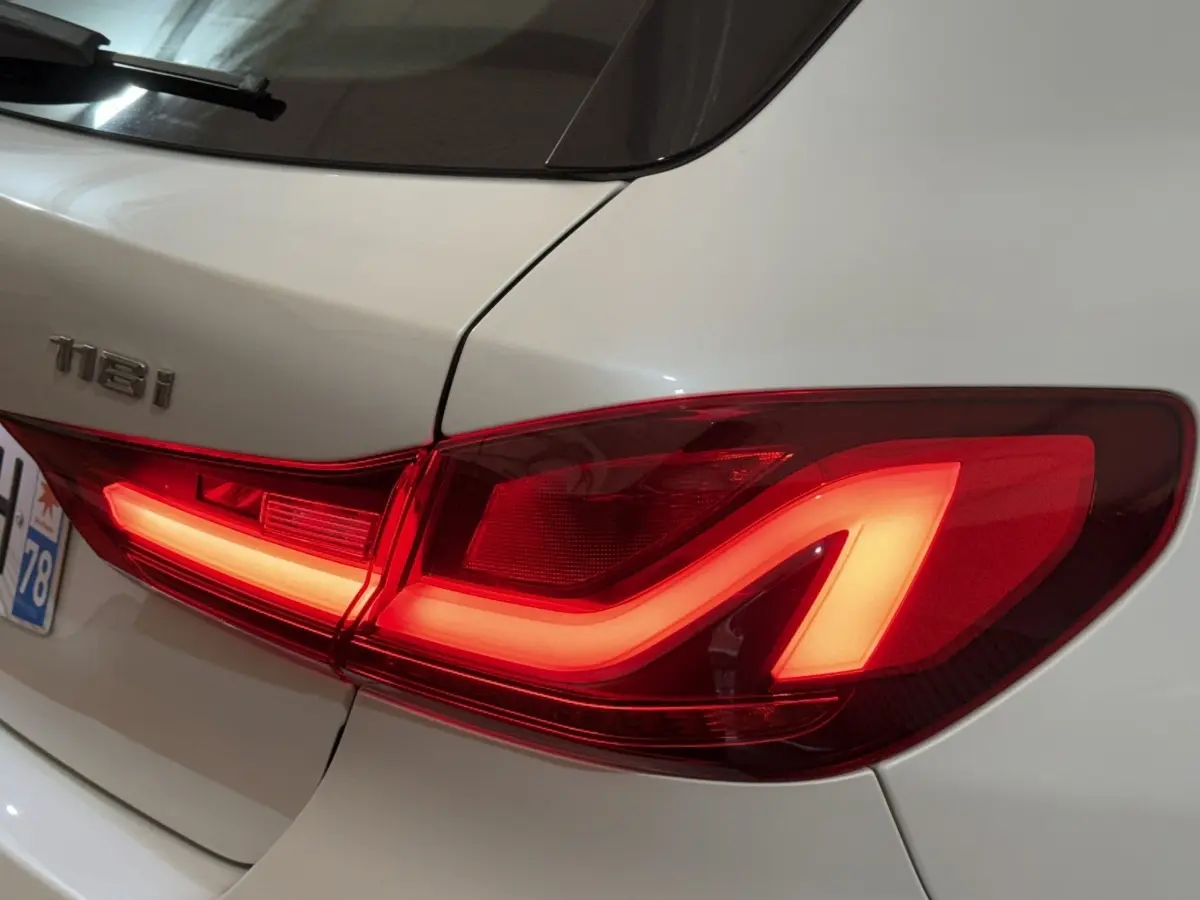 Feu arrière droit allumé sur BMW Série 1 118i blanc, vue rapprochée du coffre et du badge arrière.