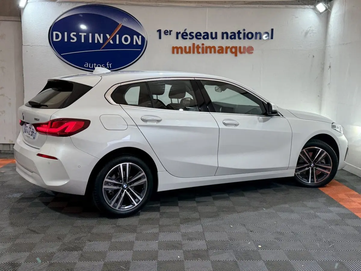 BMW Série 1 116i Business Design blanche vue de profil côté droit dans un showroom avec logo Distinxion en arrière-plan.