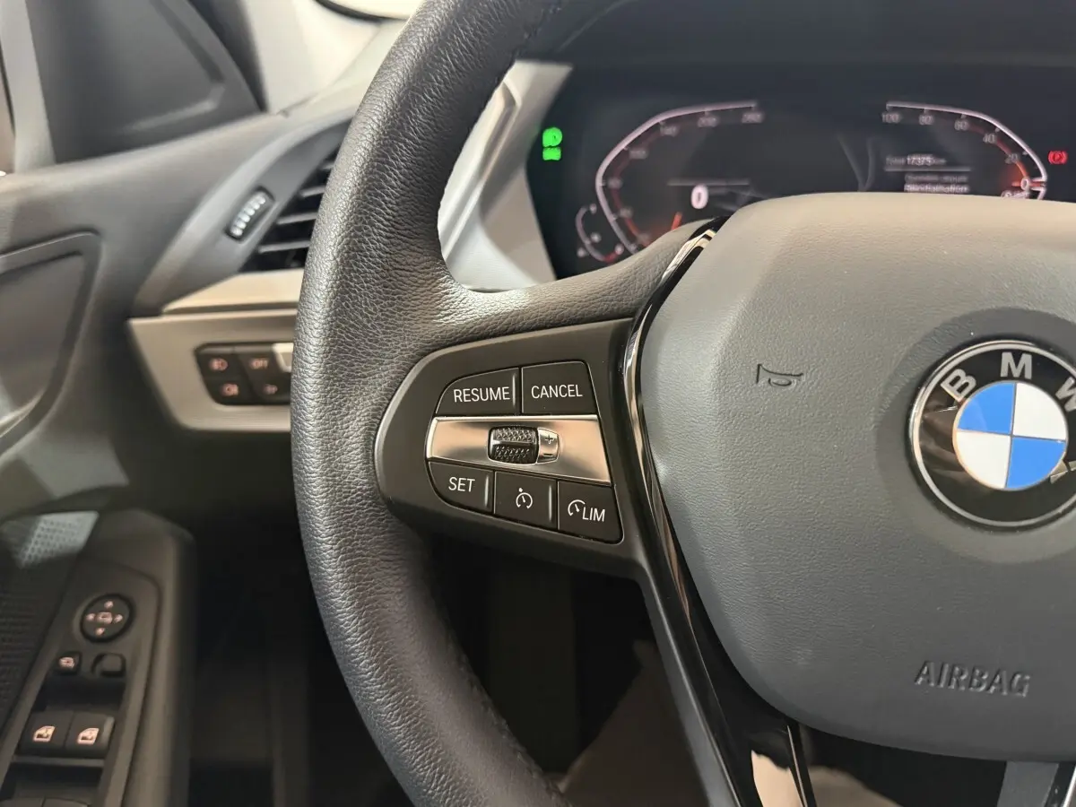 Gros plan sur le volant cuir noir de la BMW Série 1 116i 2023 avec commandes de régulateur et logo central visible.