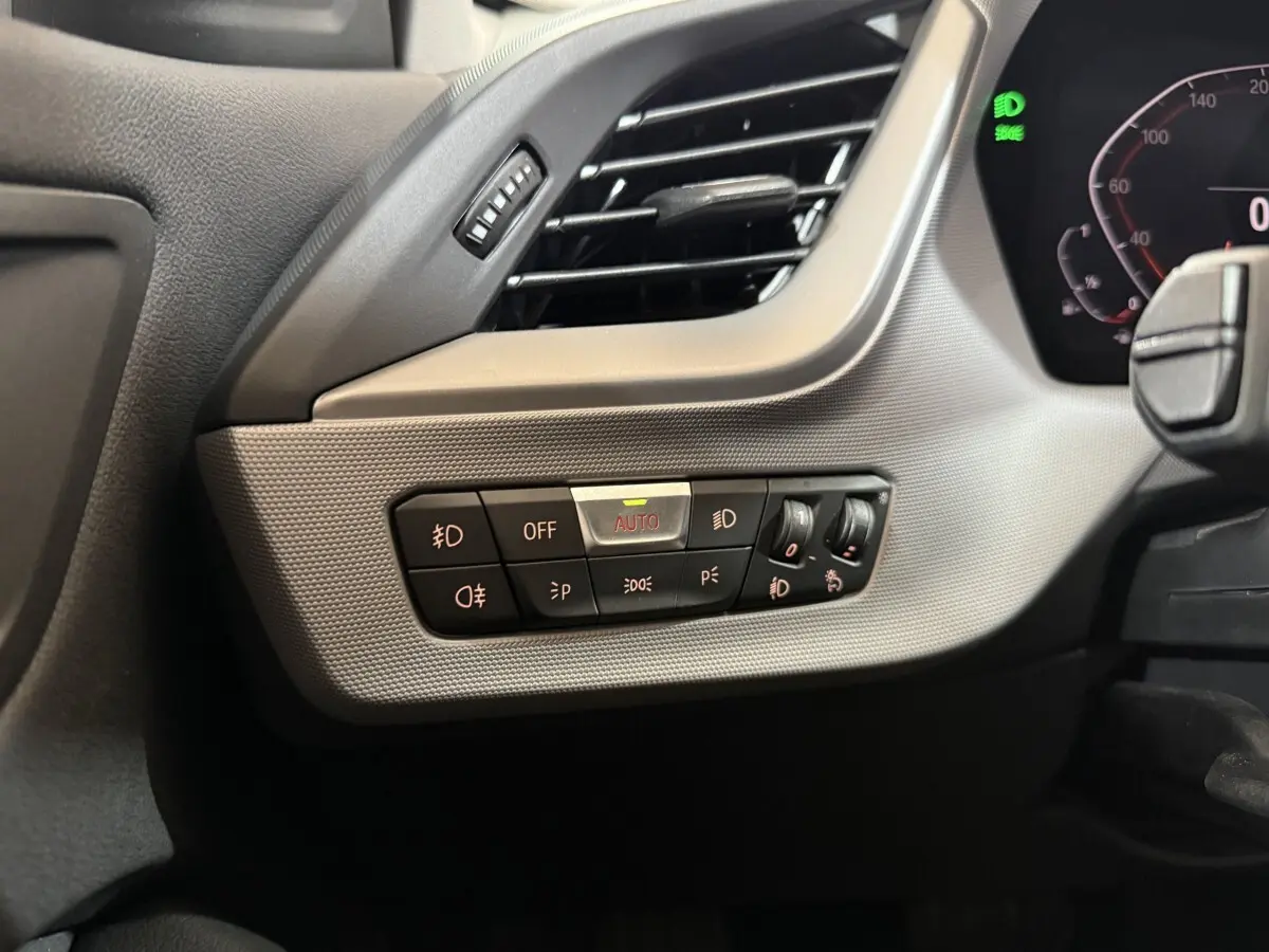 Gros plan sur le tableau de bord côté gauche d'une BMW Série 1 blanche, montrant les commandes d'éclairage avec réglage automatique.