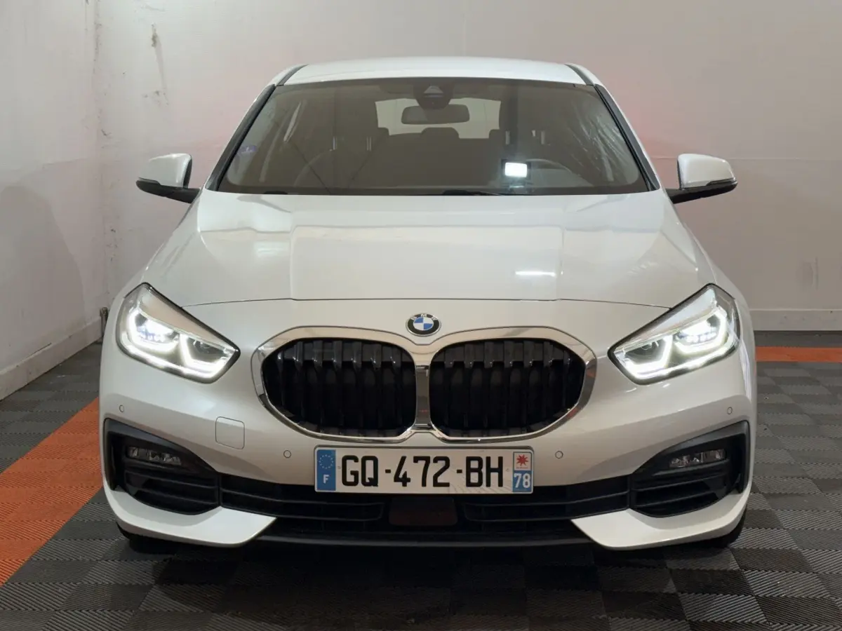 Vue frontale d'une BMW Série 1 116i blanche avec phares LED allumés et calandre noire caractéristique.