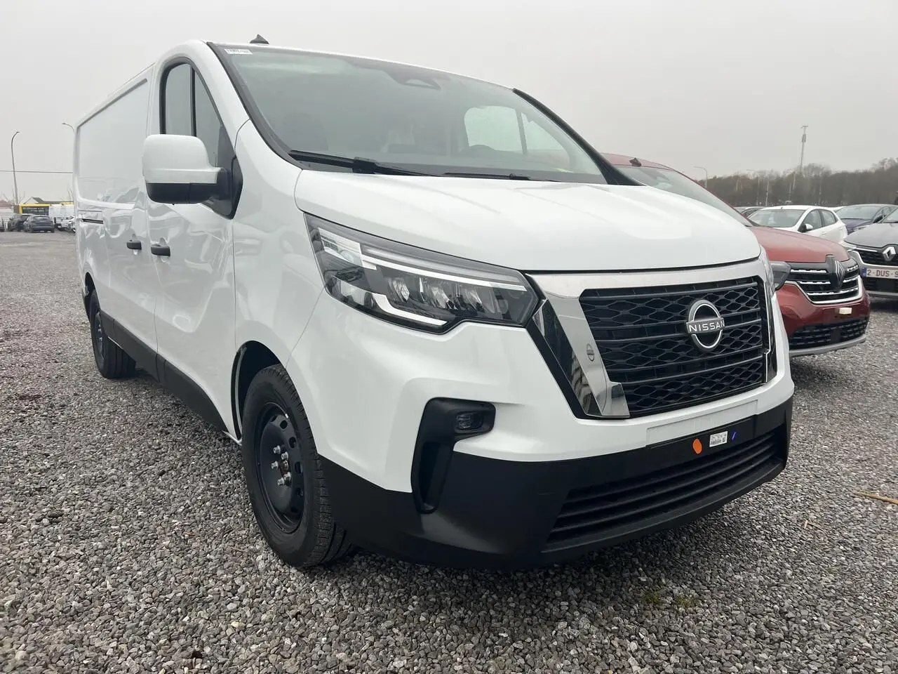 Nissan Primastar blanc en vue 3/4 avant droit sur parking extérieur avec abri métallique en arrière-plan.