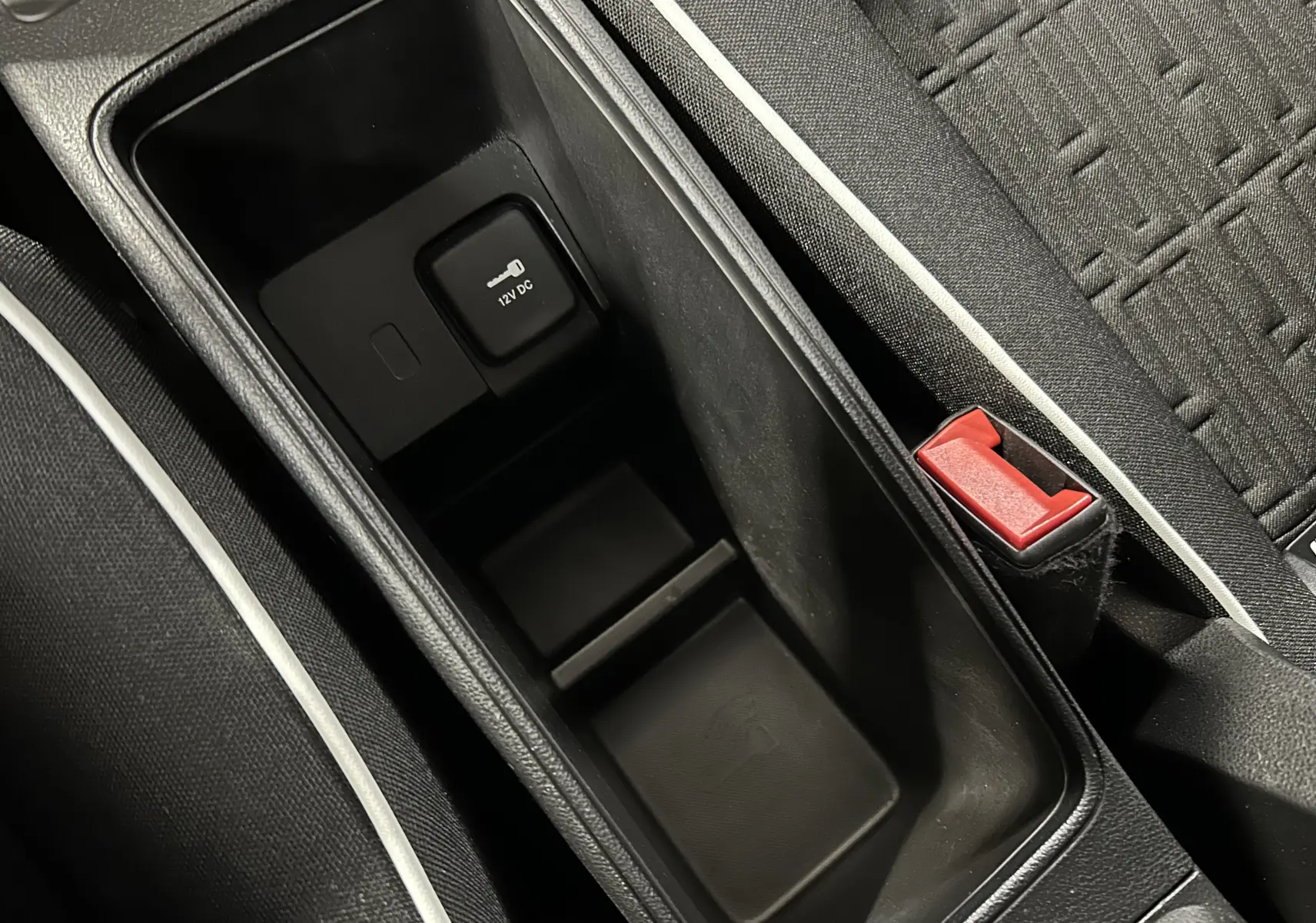 Gros plan sur la console centrale noire avec prise 12V et bouton de verrouillage, siège tissu gris avec surpiqûres blanches.