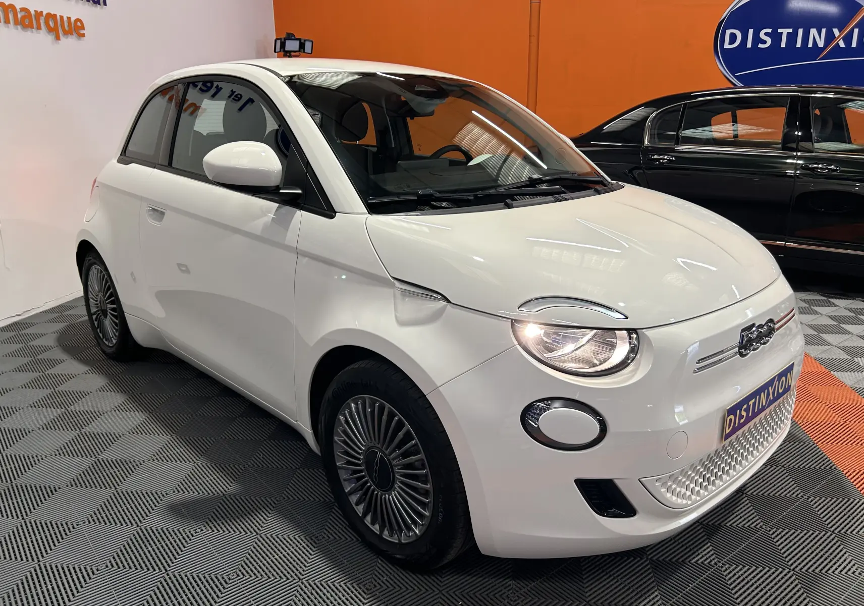 Vue 3/4 avant droite d'une FIAT 500 électrique blanche 2023 avec phares allumés et jantes stylisées.