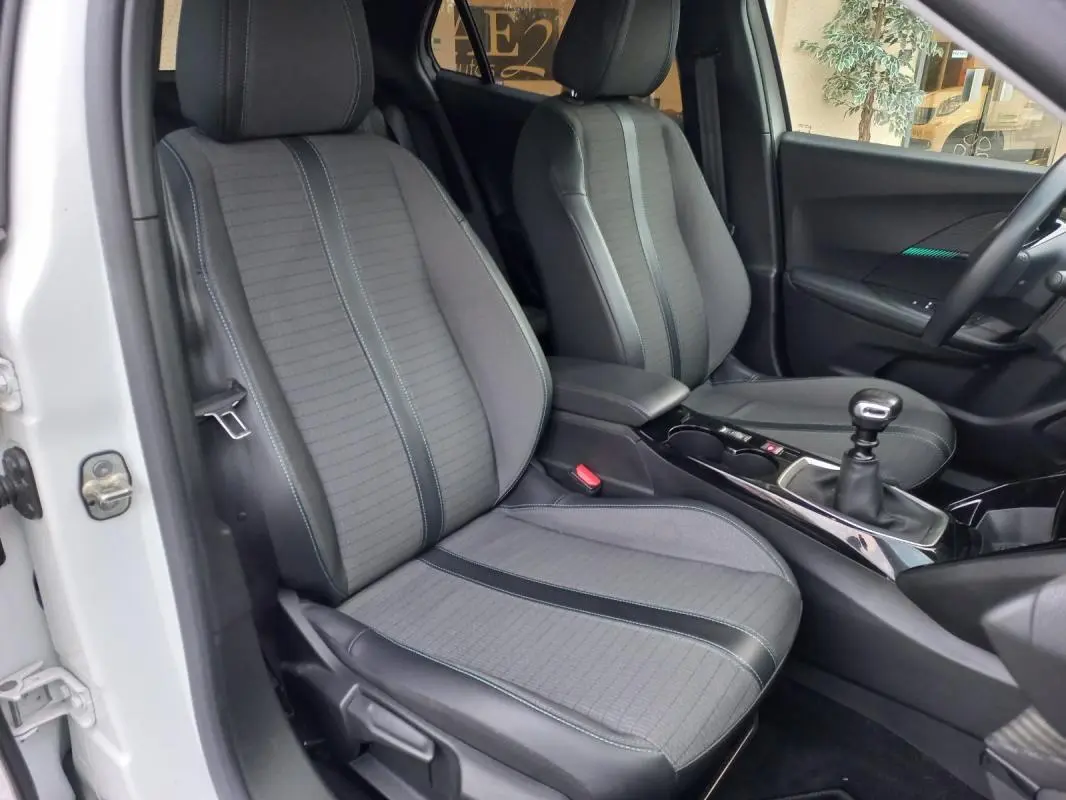Vue intérieure côté conducteur de la Peugeot 2008 blanche, montrant les sièges gris et la boîte manuelle.