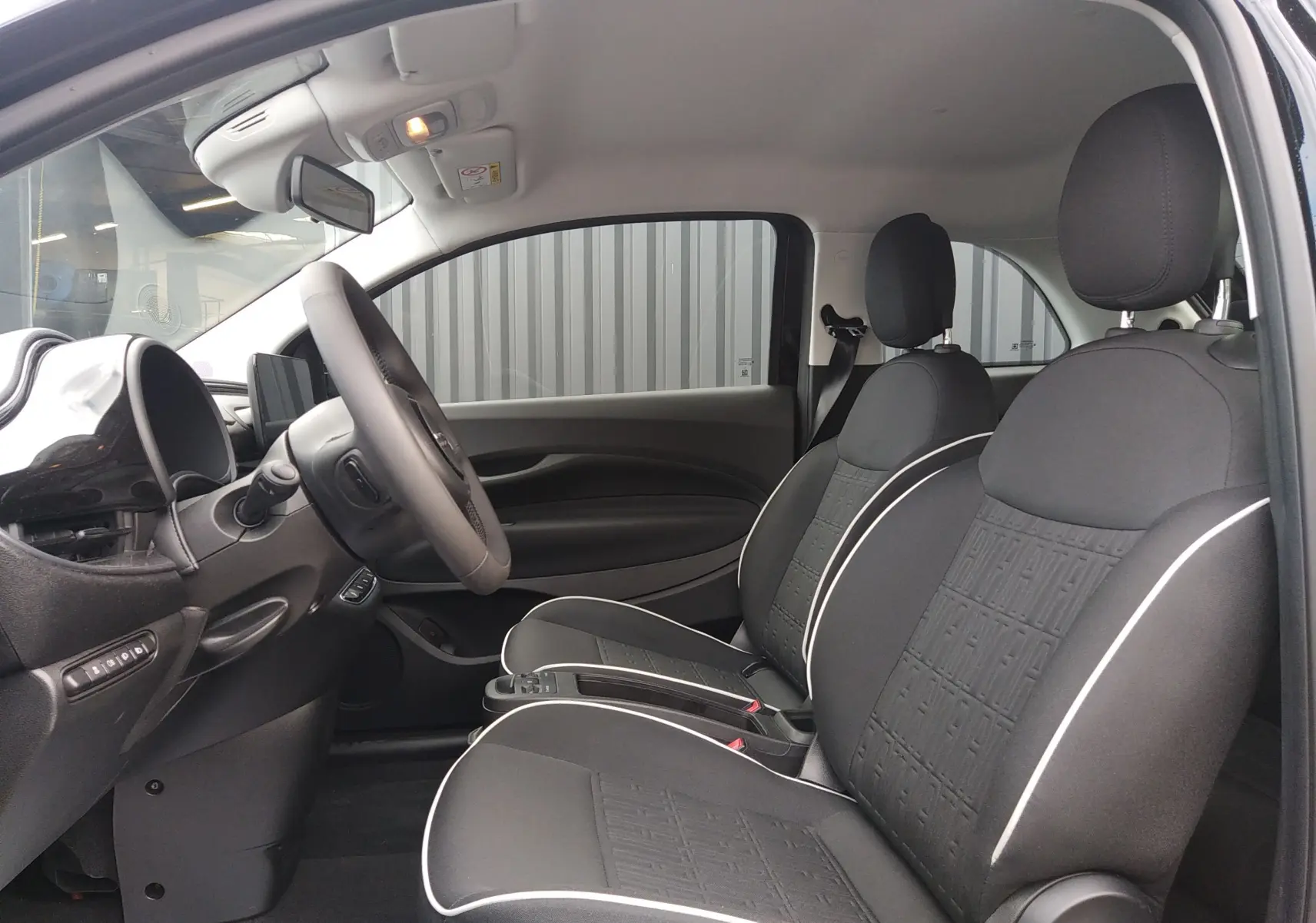 Vue intérieure côté conducteur de la Fiat 500 électrique 2024, sièges noirs avec surpiqûres blanches et volant multifonction.