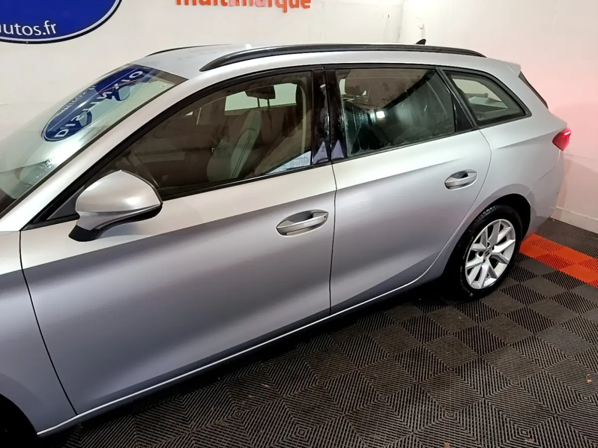 Vue latérale côté gauche d'une SEAT LEON gris clair 2022 en stationnement intérieur sur sol damier noir et orange.